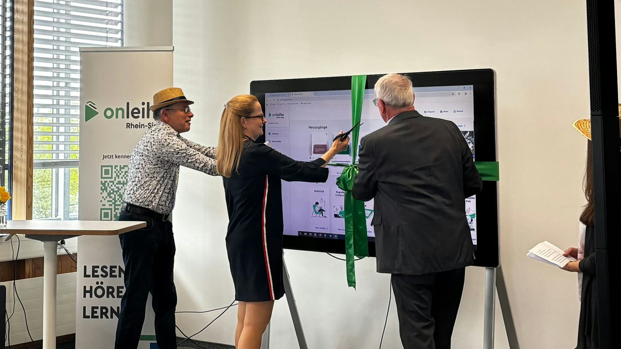 Landrat Sebastian Schuster und die Präsidentin der Hochschule Bonn-Rhein-Sieg stehen vor einem Fernseher, um den eine grüne Schleife gebunden ist und schneiden das grüne Schleifenband durch.
