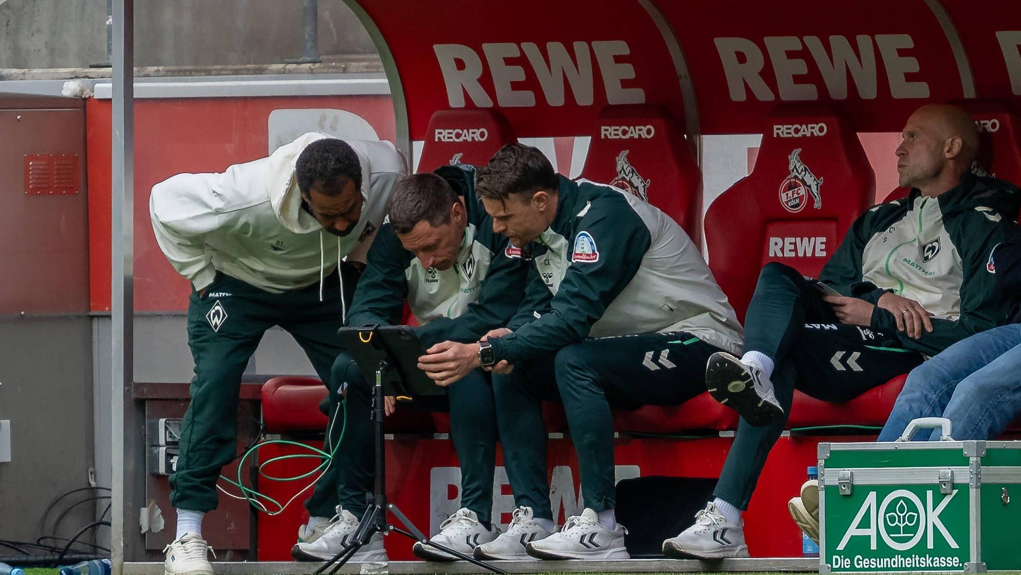 Bei einer Spielunterbrechung riskiert auch Trainer Daniel Thioune (l.) von Werder Bremen einen Blick auf das Tablet vor seiner Trainerbank.