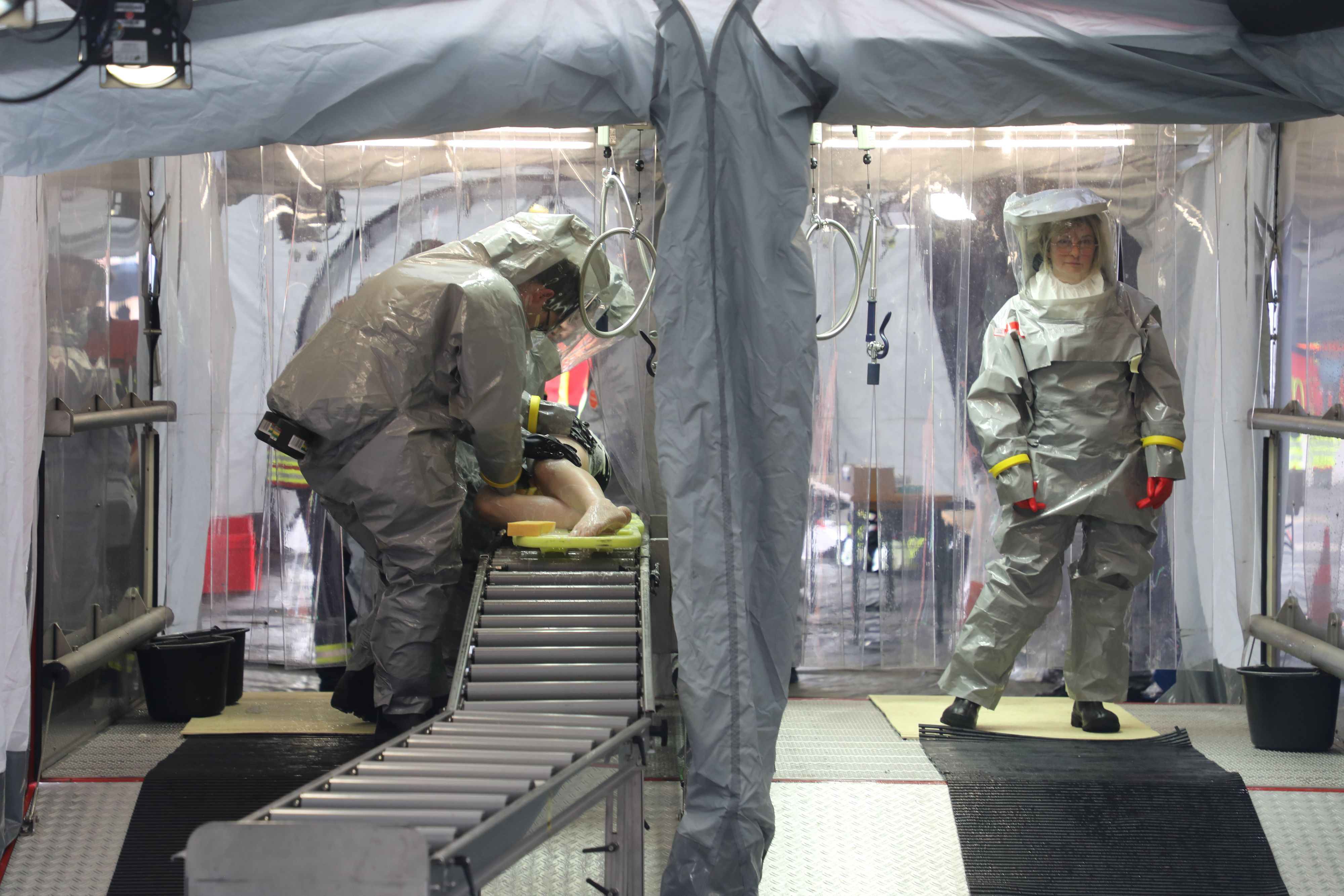 Einsatzkräfte in Schutzkleidung stehen in einem Dekontaminations-Abrollcontainer und duschen kontaminierte Patienten ab.