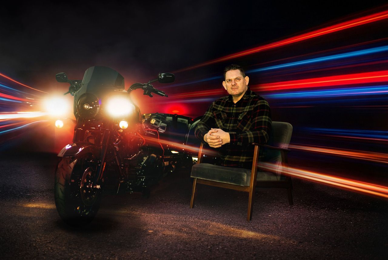 Das Foto zeigt einen Mann, der keine Beine mehr besitzt, auf einem Stuhl. Neben ihm steht ein Motorrad.