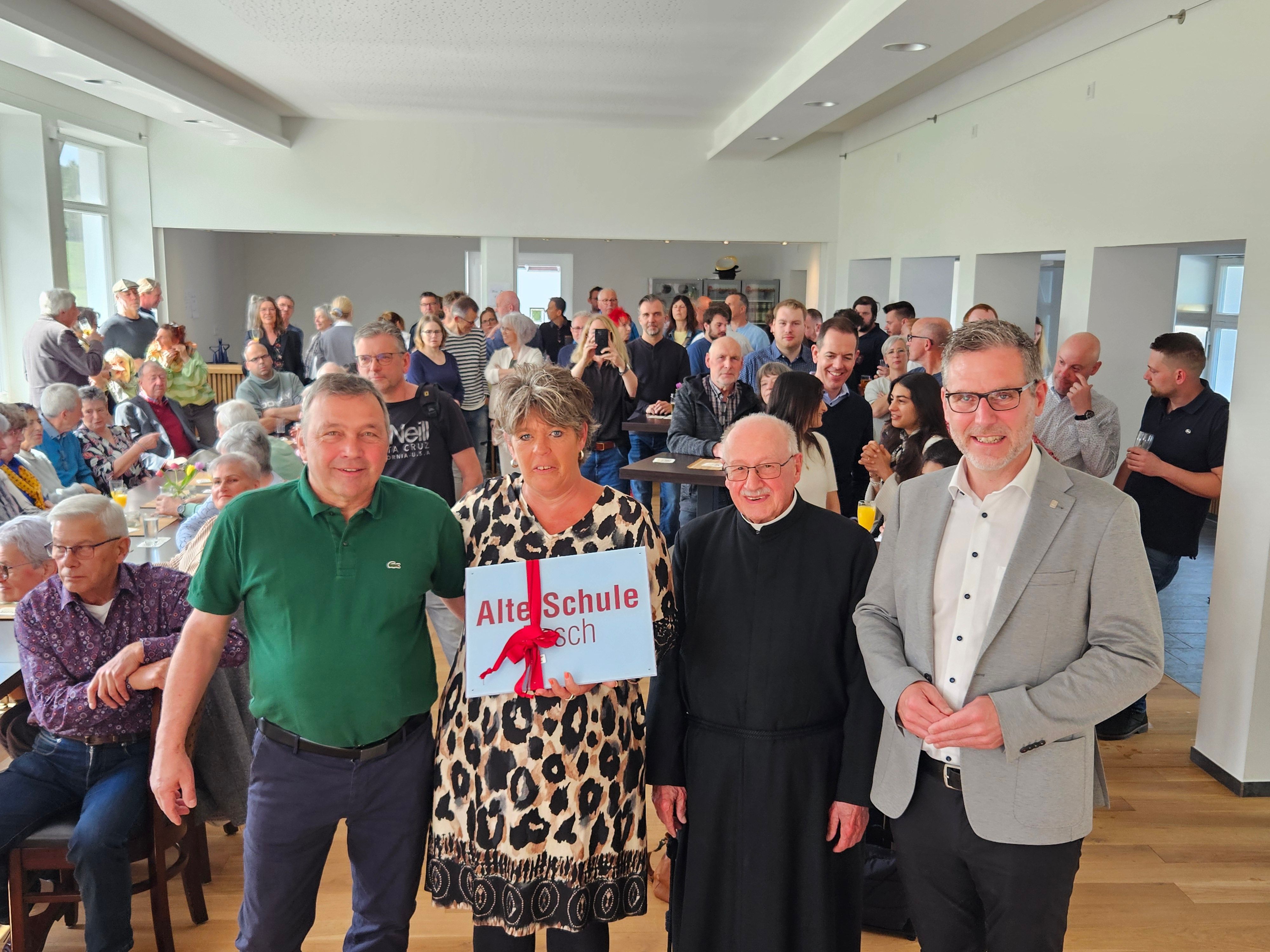 Drei Männer und eine Frau stehen nebeneinander. Die Frau hält ein Schild, auf dem Alte Schule Pesch steht. Im Hintergrund sind die Gäste der Einweihung zu sehen.