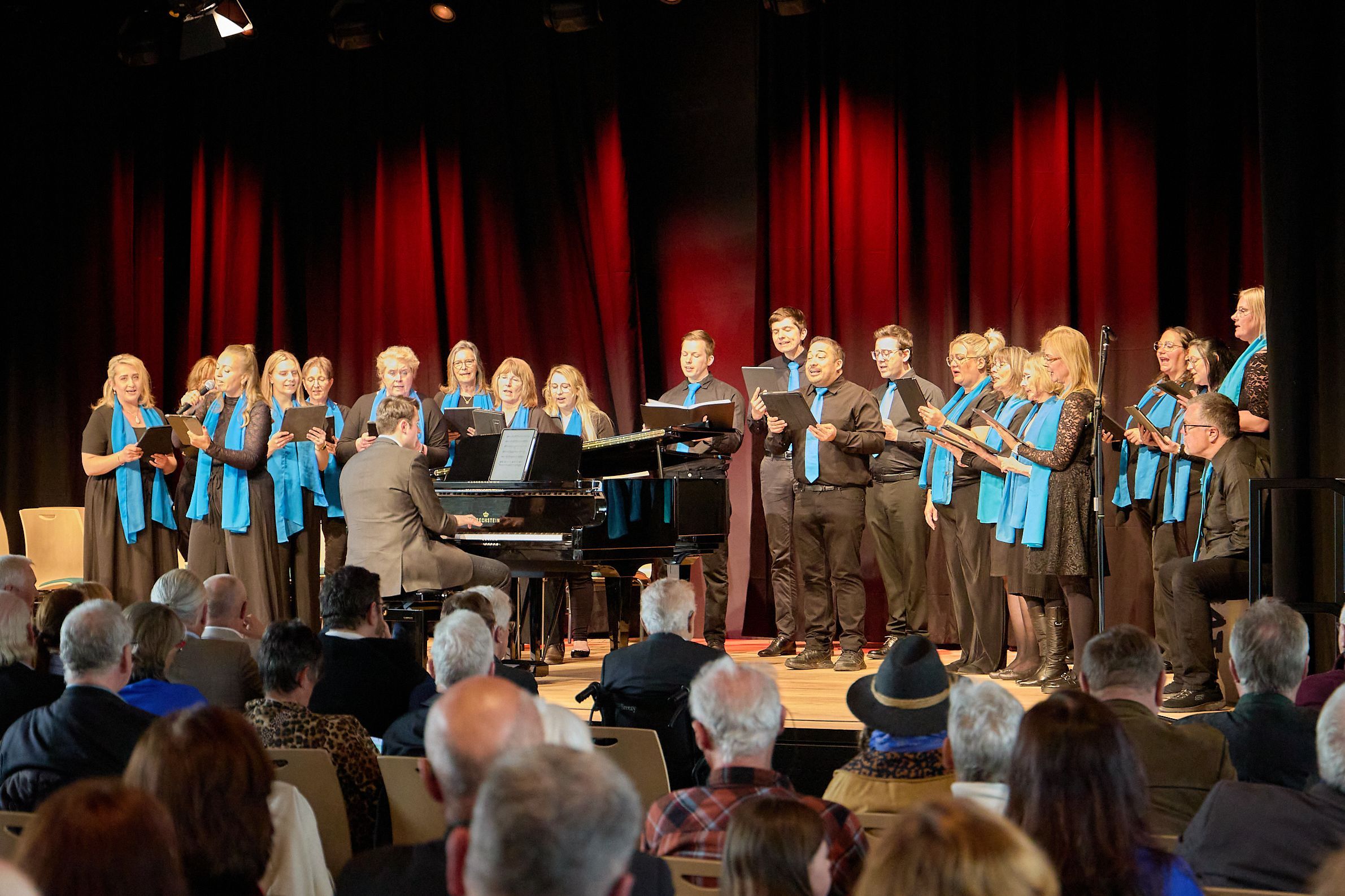 Das Bild zeigt den Chor auf der Bühne mit einem Pianisten.