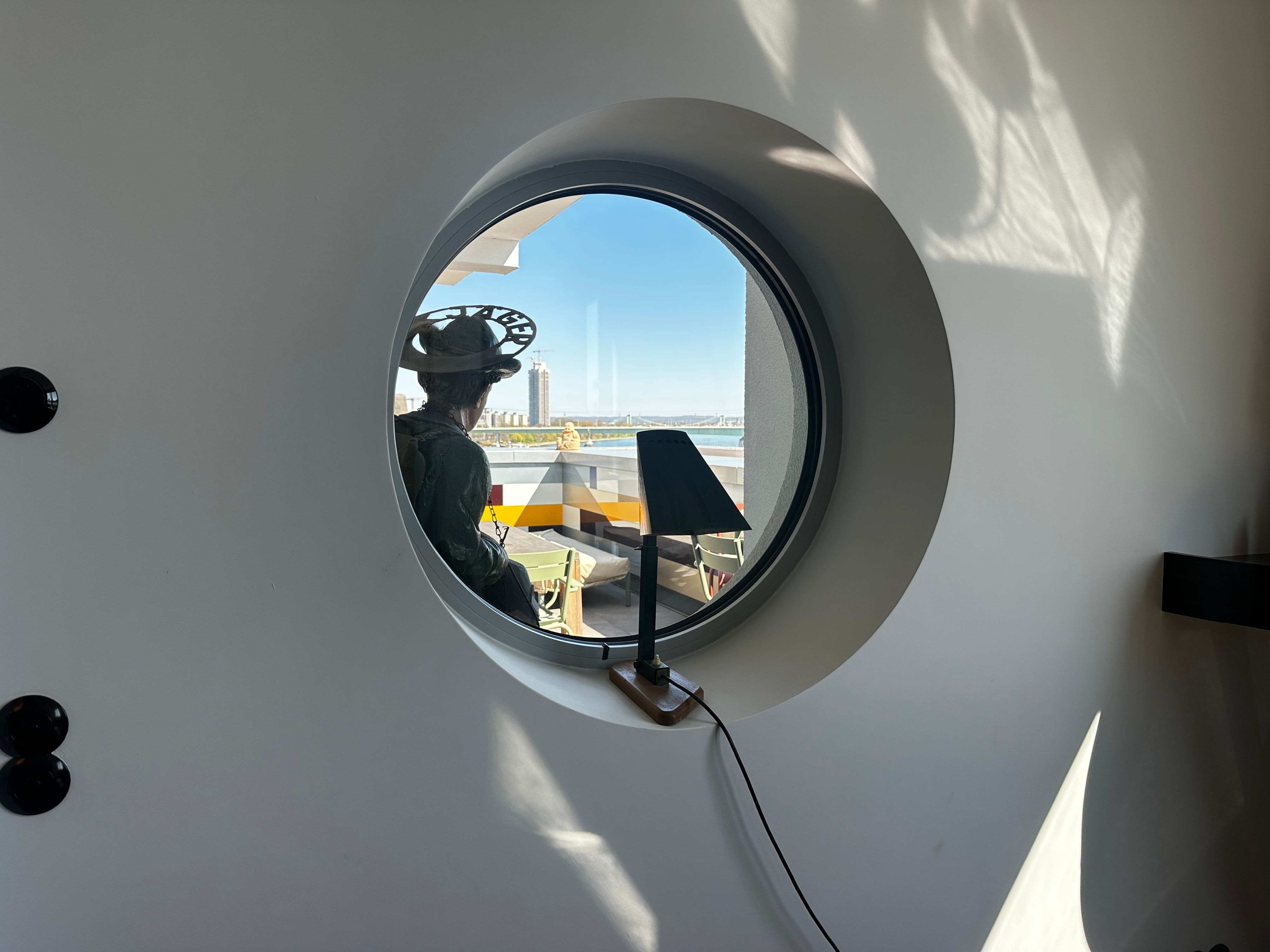 Ein rundes Fenster gibt den Blick frei auf einen Balkon, dahinter der Rhein mit dem Messeturm.
