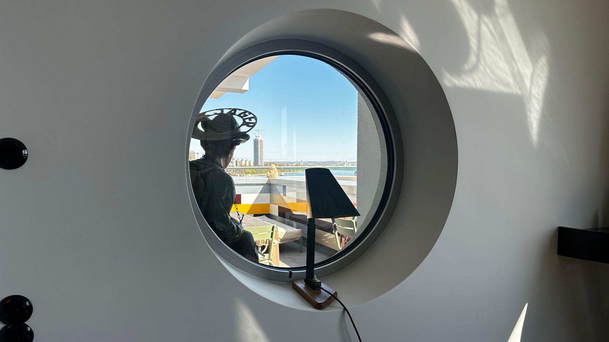 Ein rundes Fenster gibt den Blick frei auf einen Balkon, dahinter der Rhein mit dem Messeturm.