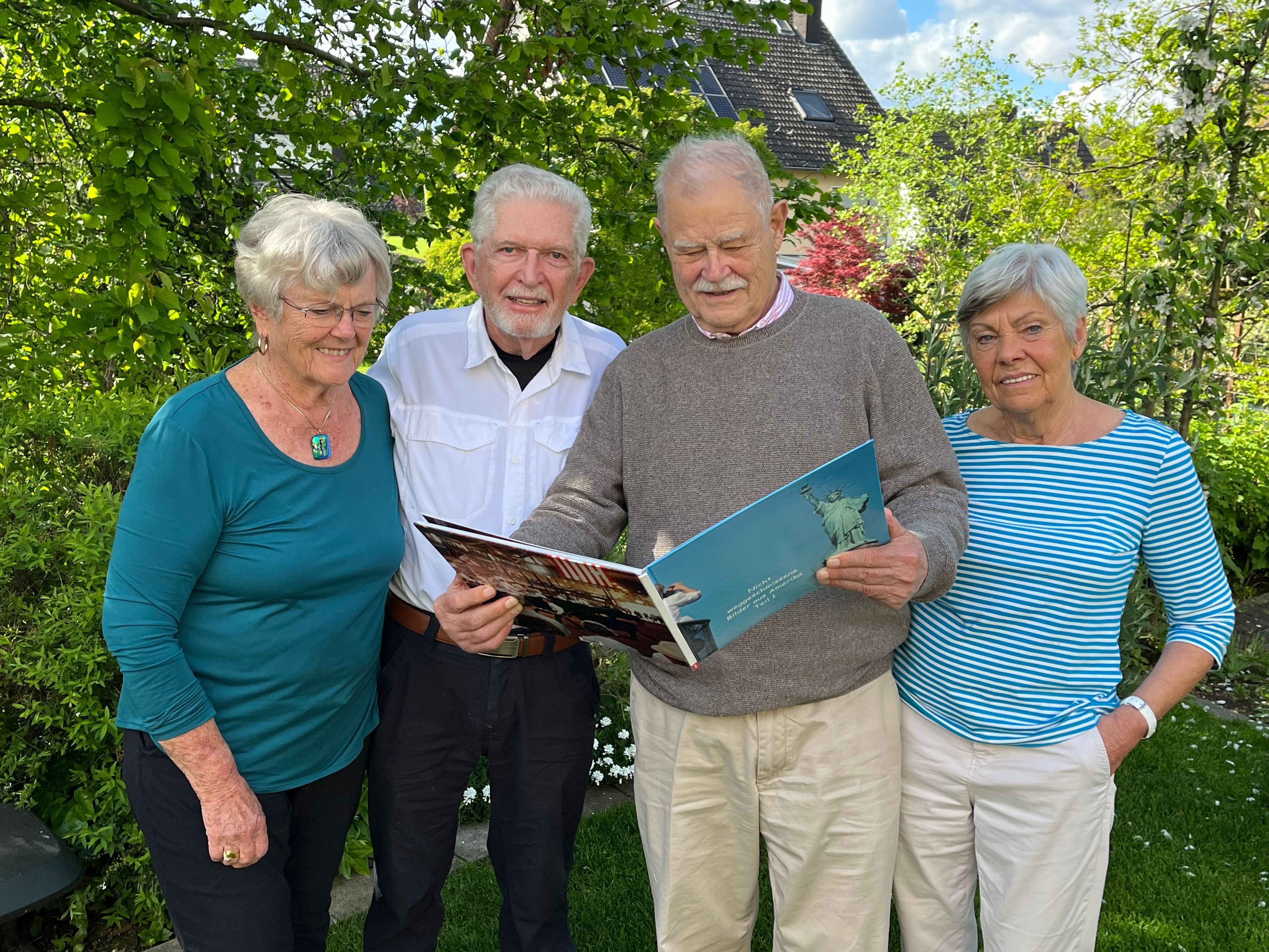 Zwei Frauen und zwei Männer stehen in einem Garten vor grünen Büschen und Bäumen. Einer der Männer hält ein Fotobuch.