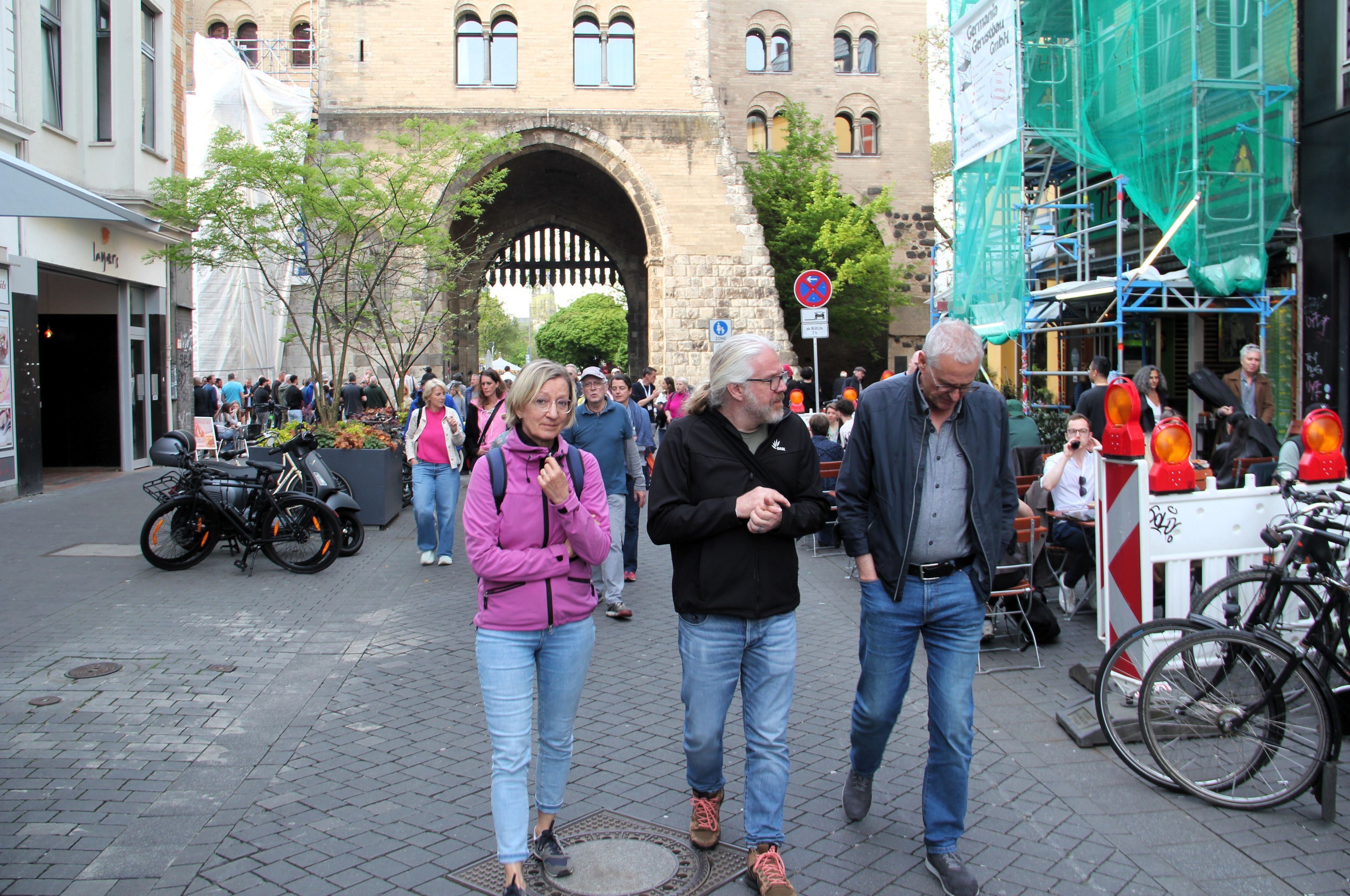 Drei Menschen laufen über den Eigelstein, im Hintergrund ist die Eigelsteintorburg sehen.