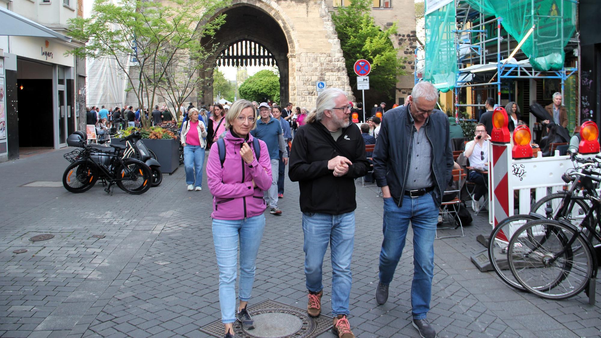 Drei Menschen laufen über den Eigelstein, im Hintergrund ist die Eigelsteintorburg sehen.