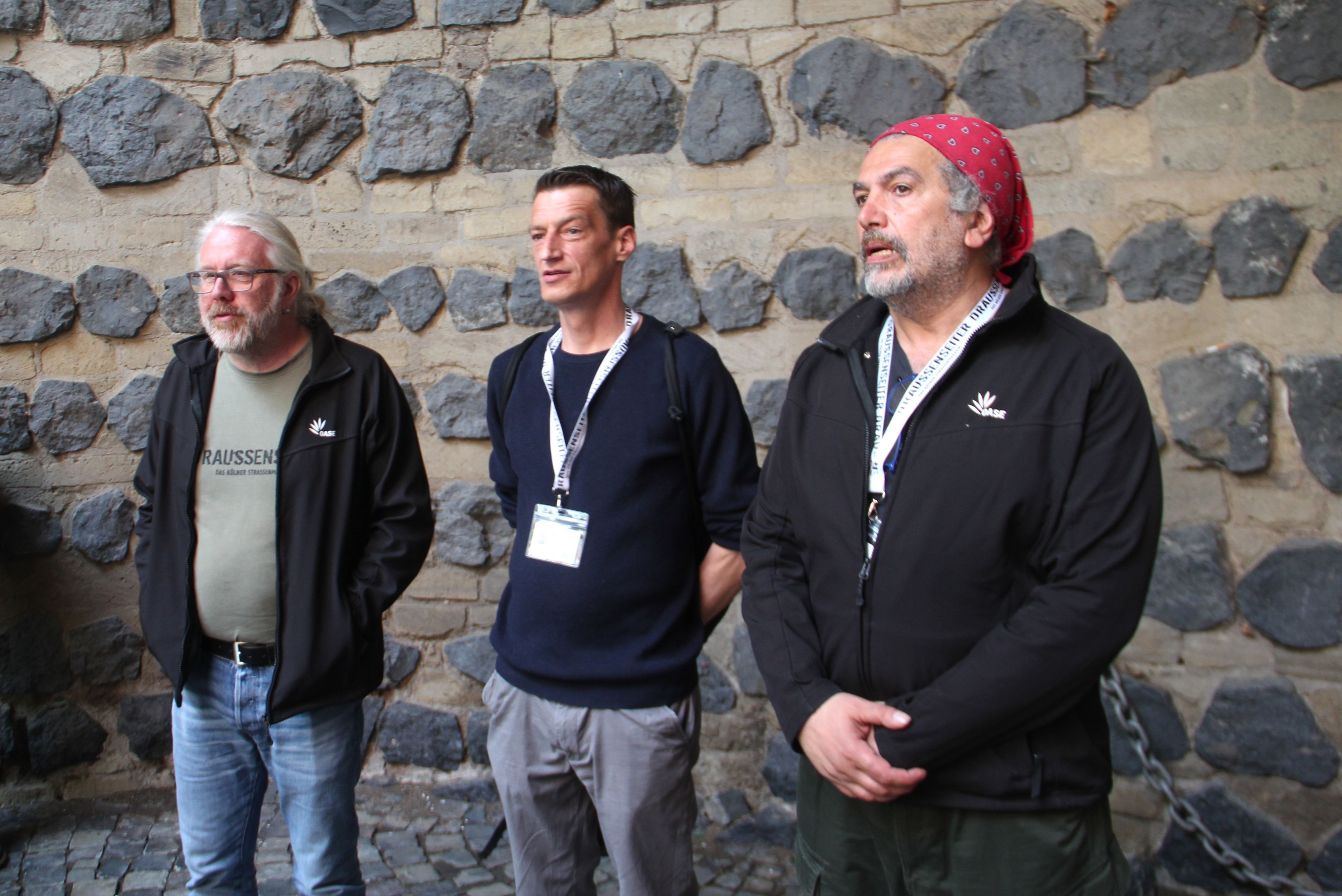 Drei Menschen stehen vor einer Mauer mit groben Steinen.