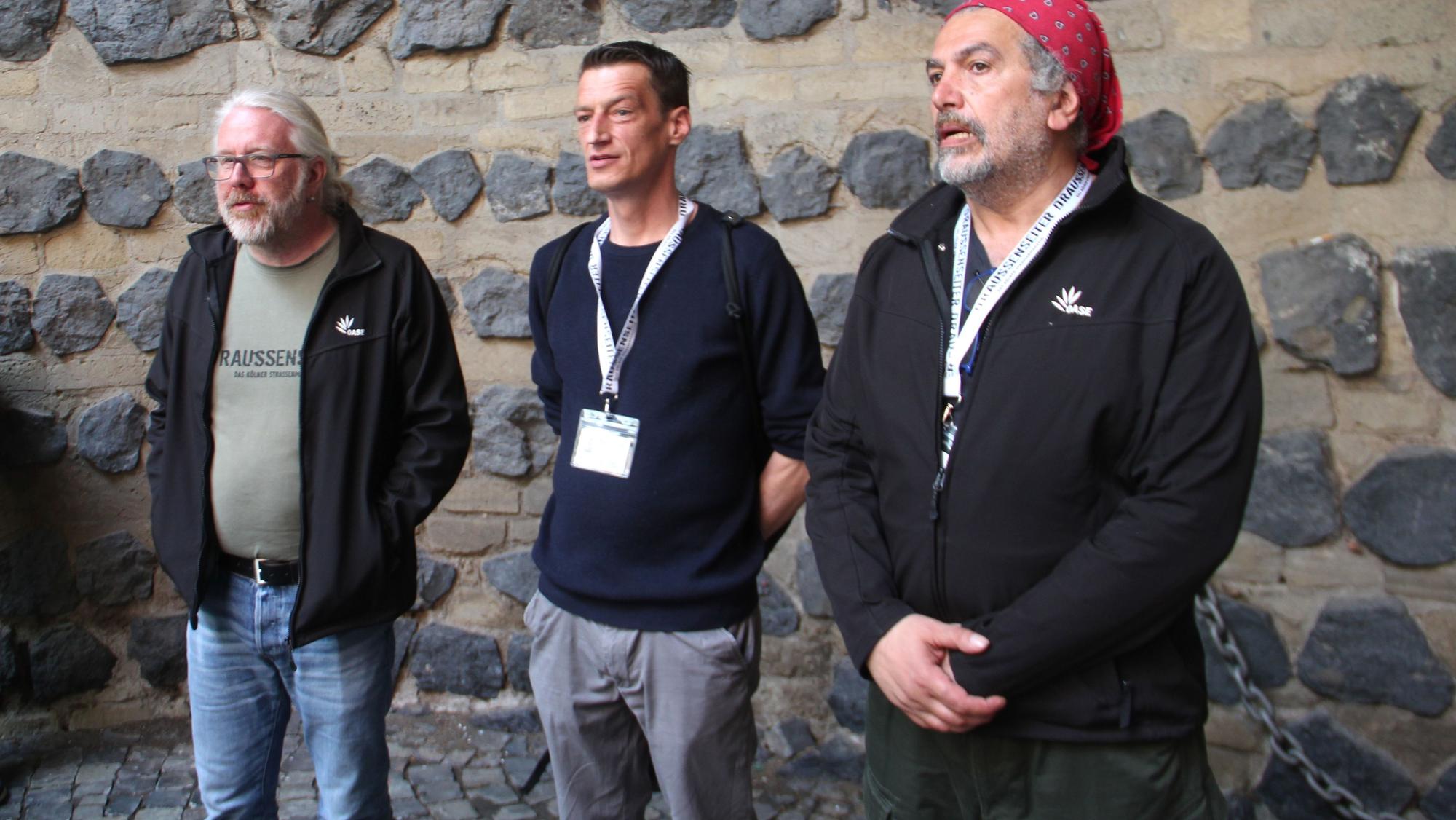 Drei Menschen stehen vor einer Mauer mit groben Steinen.