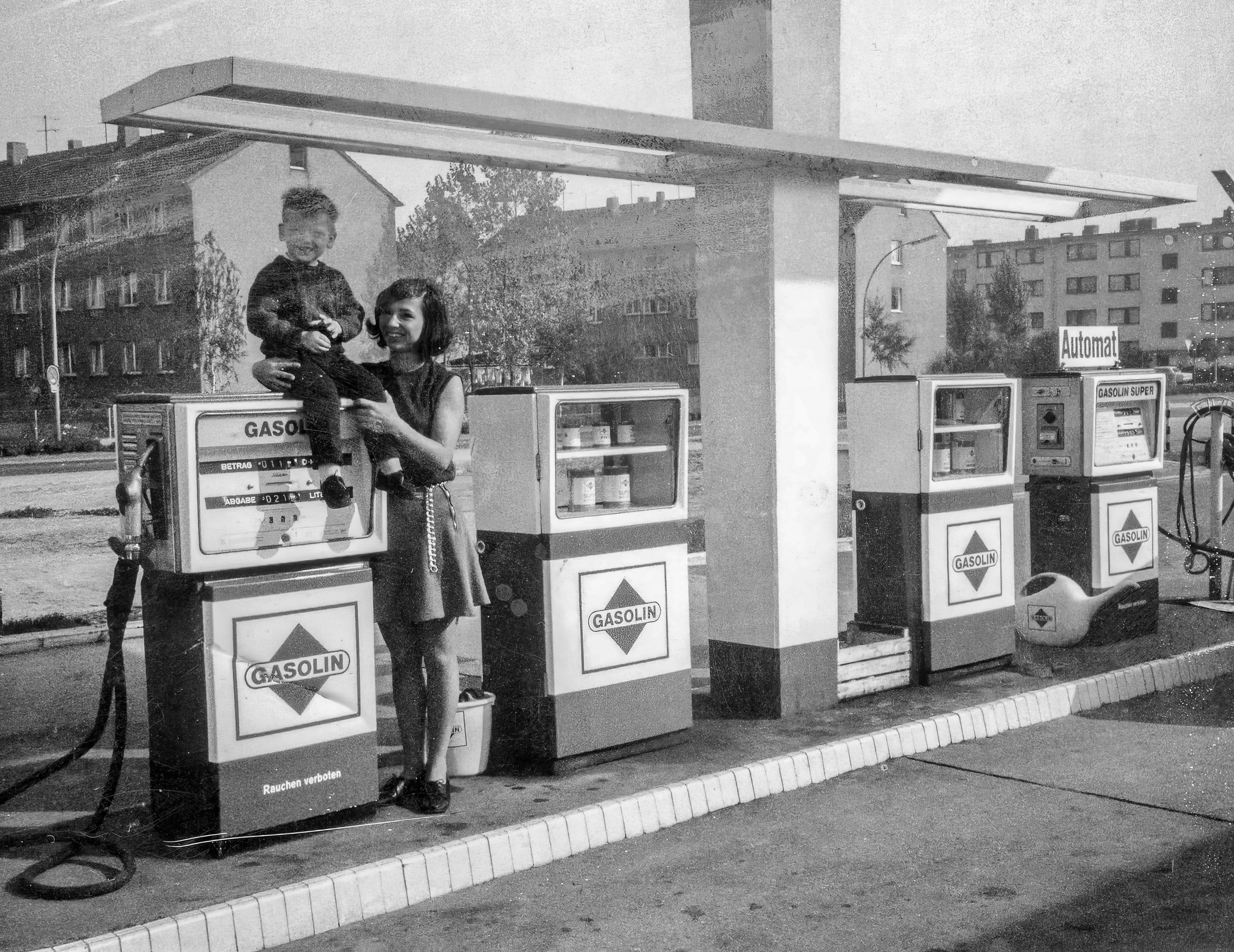 Ein Schwarz-Weiß-Foto zeigt einen kleinen Jungen, der auf einer Zapfsäule sitzt, eine Frau mit dunklem Haar hält ihn fest.