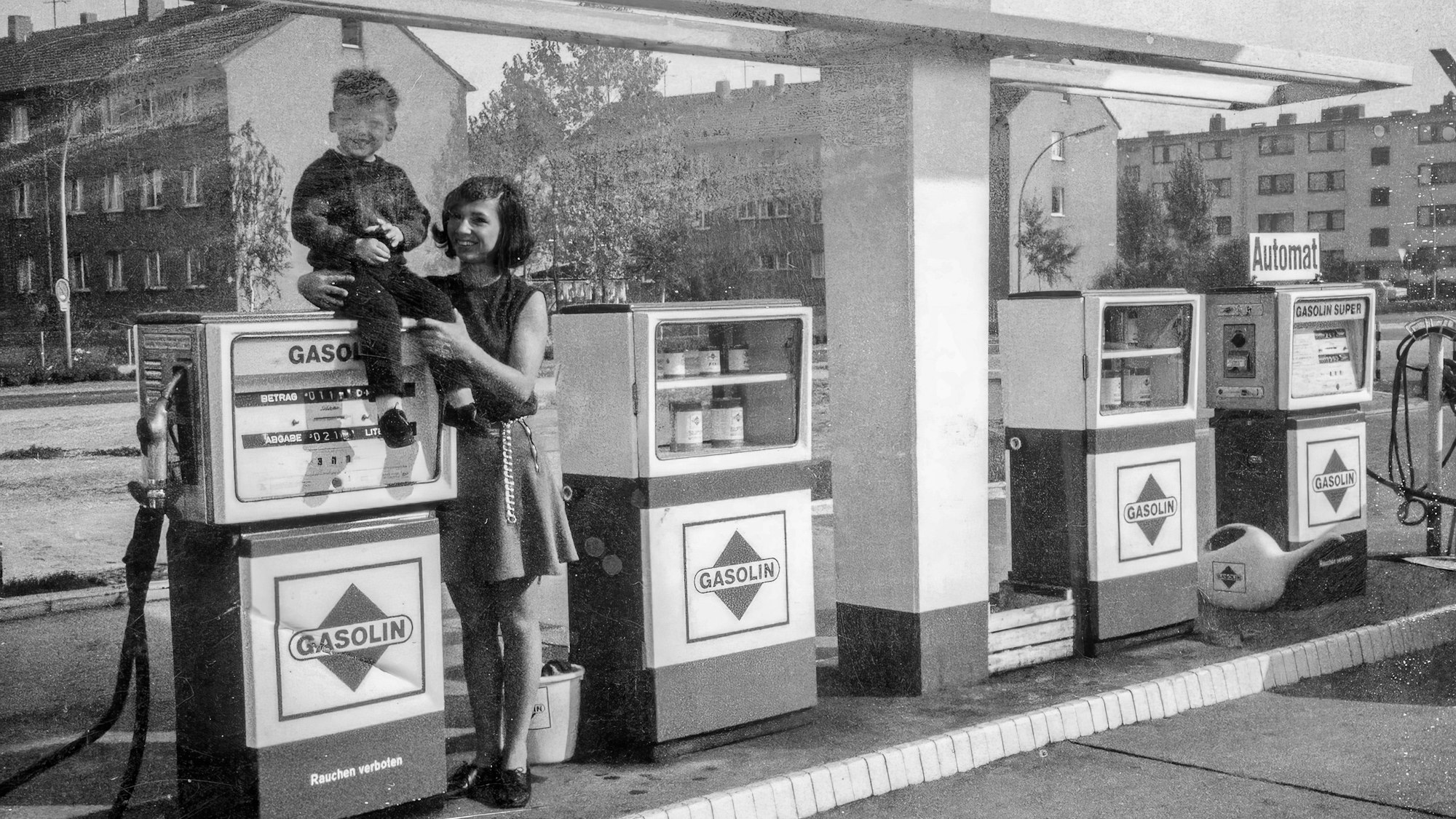 Ein Schwarz-Weiß-Foto zeigt einen kleinen Jungen, der auf einer Zapfsäule sitzt, eine Frau mit dunklem Haar hält ihn fest.