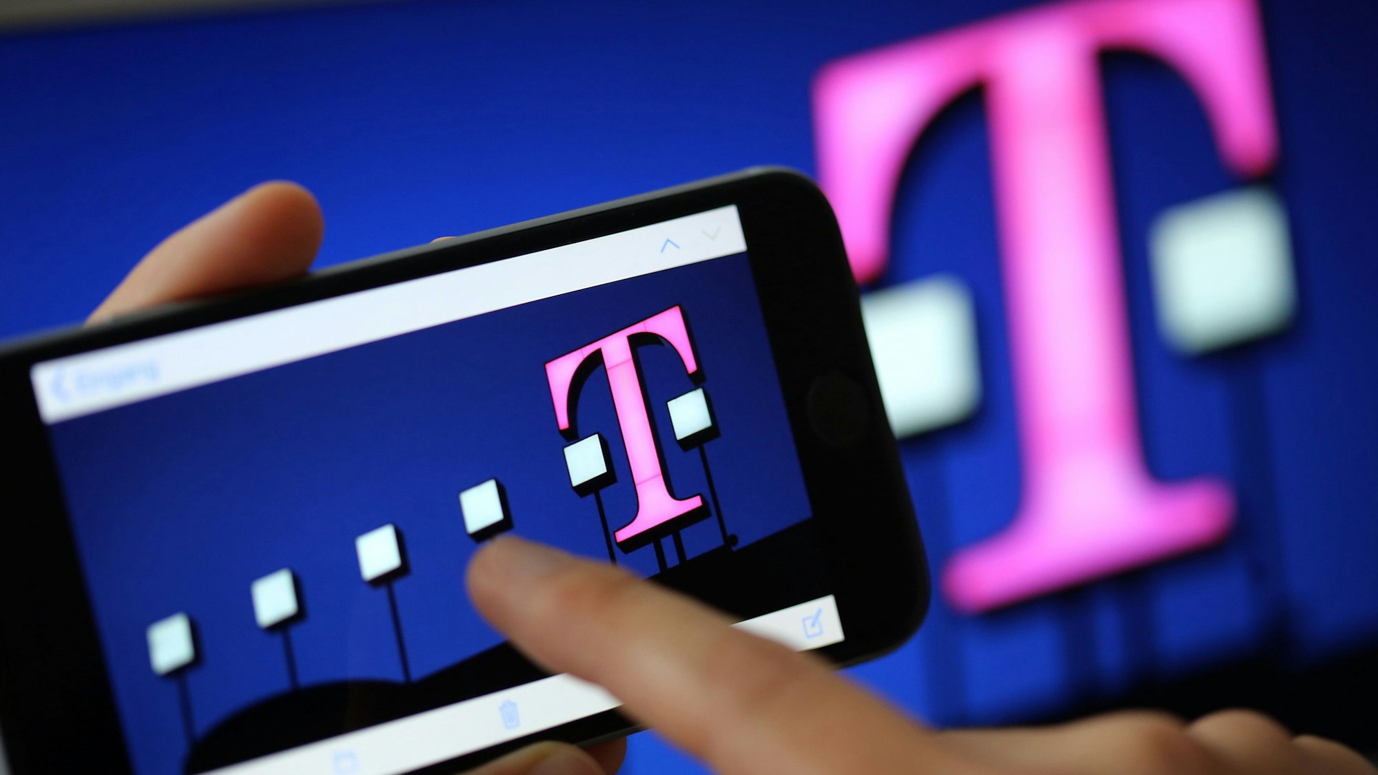 Ein Frau tippt auf ihr Smartphone, auf dem ein Telekom-Logo zu sehen ist. Bei einer Tochterfirma des Konzerns gab es jetzt Durchsuchungen. (Symbolbild)
