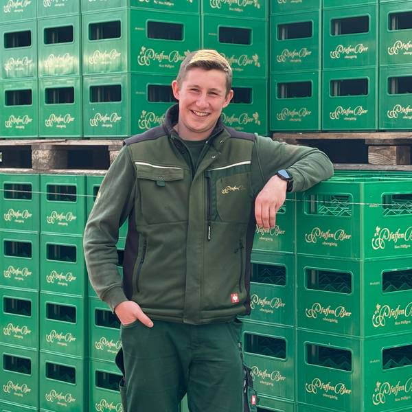 Max Päffgen lehnt mit dem linken Arm an aufgetürmten Bierkästen und lächelt in die Kamera.