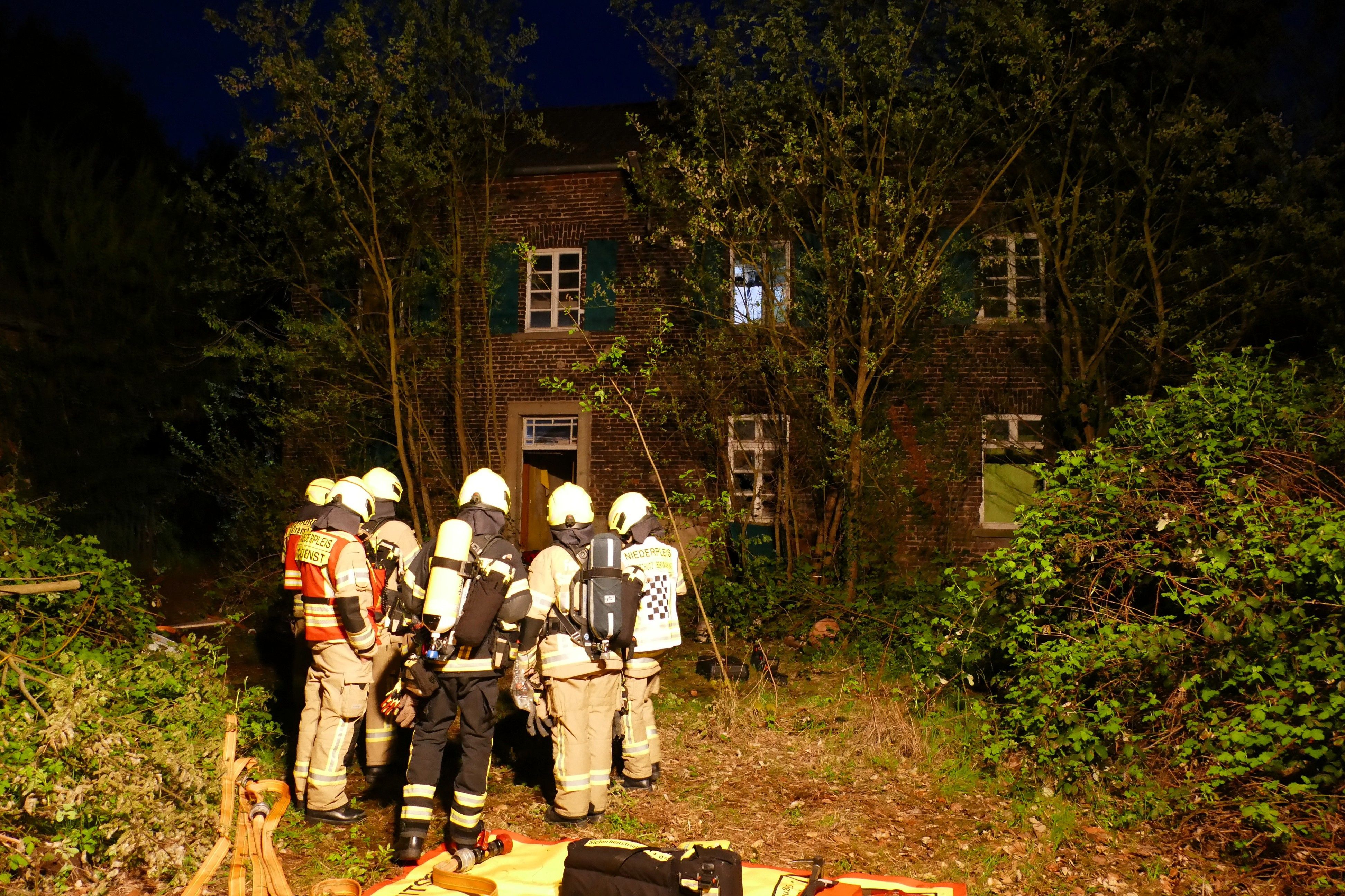 Feuerwehrleute stehen vor dem leerstehenden Haus auf einem zugewucherten Grundstück.