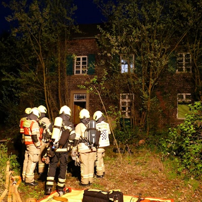 Feuerwehrleute stehen vor dem leerstehenden Haus auf einem zugewucherten Grundstück.
