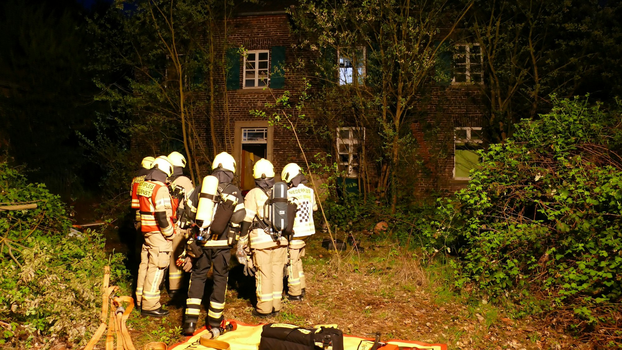 Feuerwehrleute stehen vor dem leerstehenden Haus auf einem zugewucherten Grundstück.