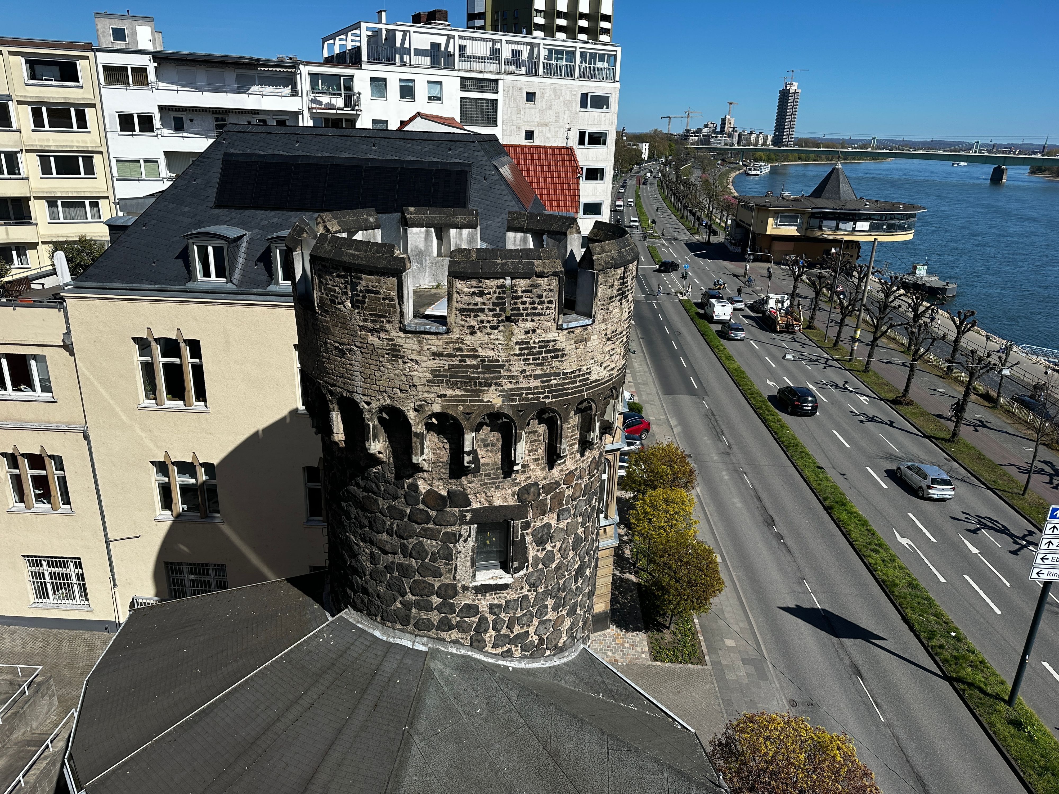 Von einem Balkon aus fällt der Blick auf einen steinernen Turm, eine vierspurige Straße, die Bastei, den Rhein, die Mülheimer Brücke und ein Hochhaus.