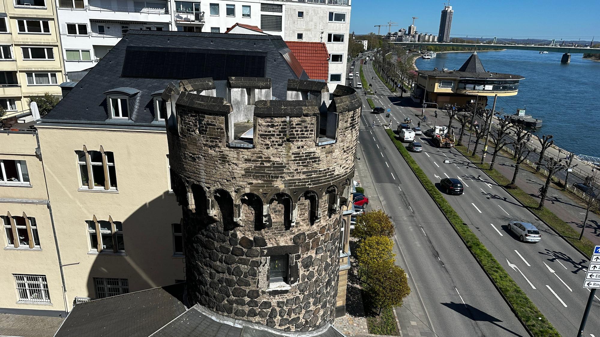 Von einem Balkon aus fällt der Blick auf einen steinernen Turm, eine vierspurige Straße, die Bastei, den Rhein, die Mülheimer Brücke und ein Hochhaus.