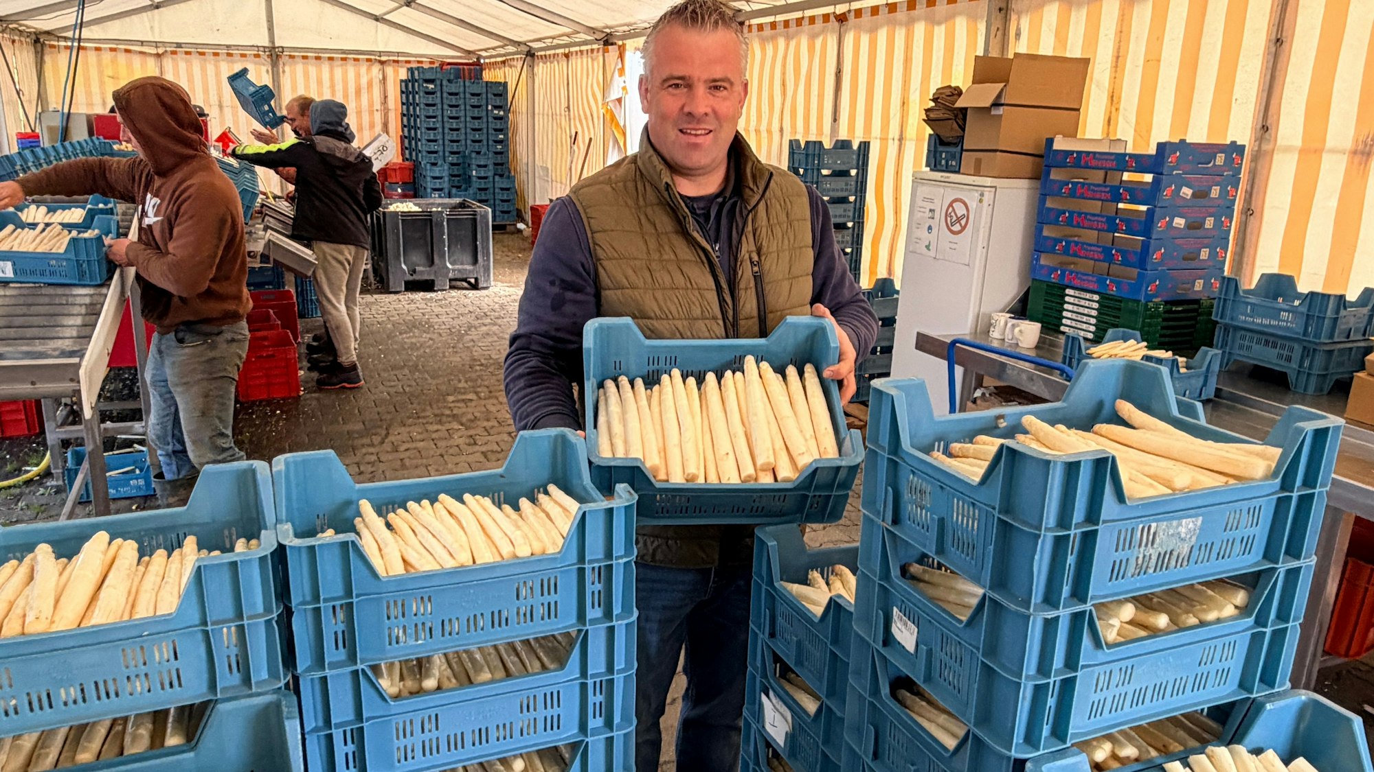 Der Landwirt steht vor den aufgestapelten Boxen, in denen der frische Spargel verkauftsfertig liegt.