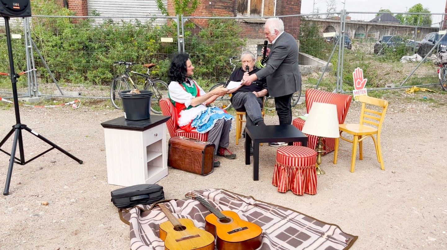 Drei Männer sind in einer Theaterszene zu sehen. Einer mit langer dunkler Perücke bekommt von einem anderen einen Vertrag zur Unterzeichnung überreicht.