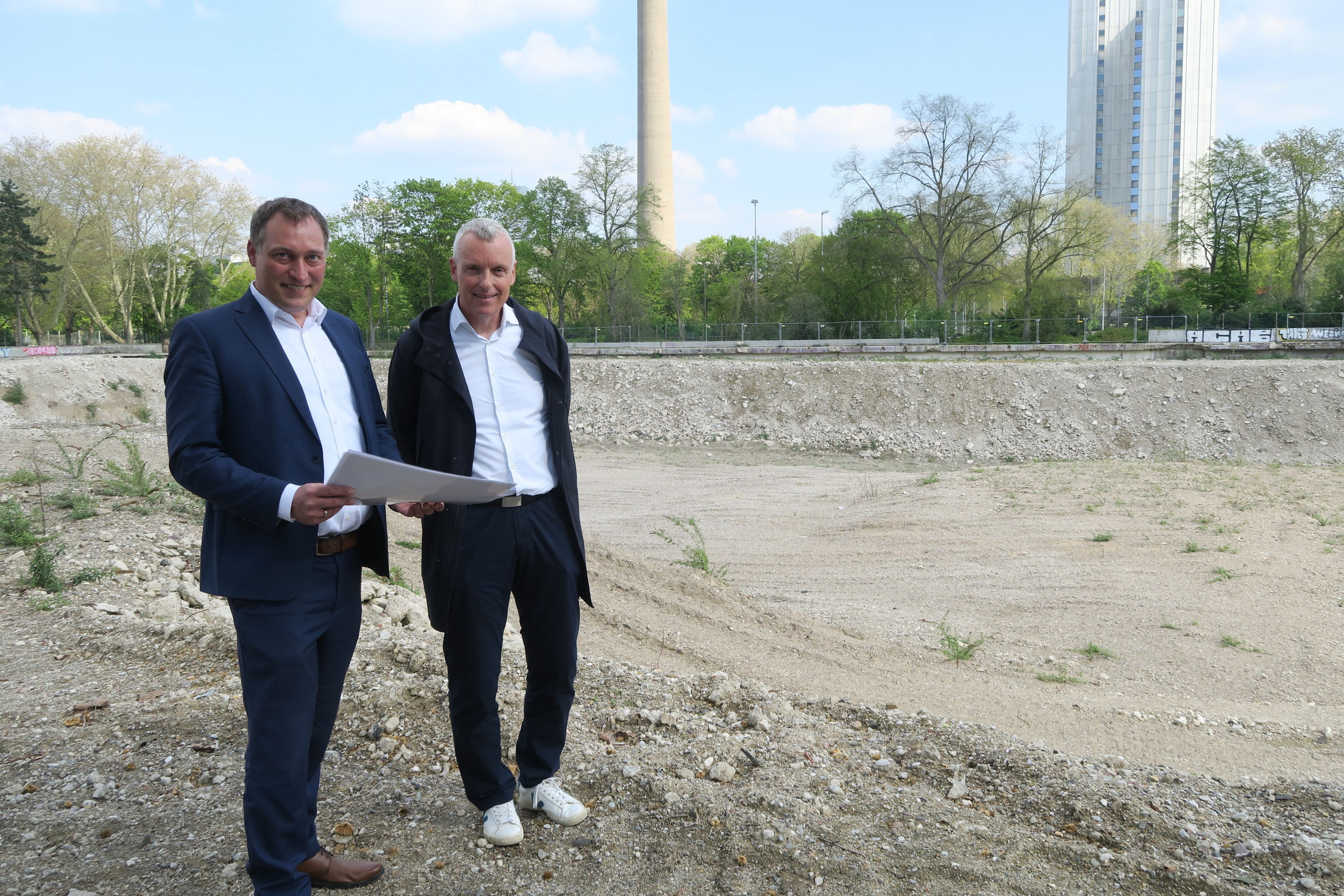 Zwei Männer in Anzügen stehen auf einem leeren Grundstück. Im Hintergrund sind der Colonius und das Telekom-Hochhaus an der Inneren Kanalstraße zu sehen.