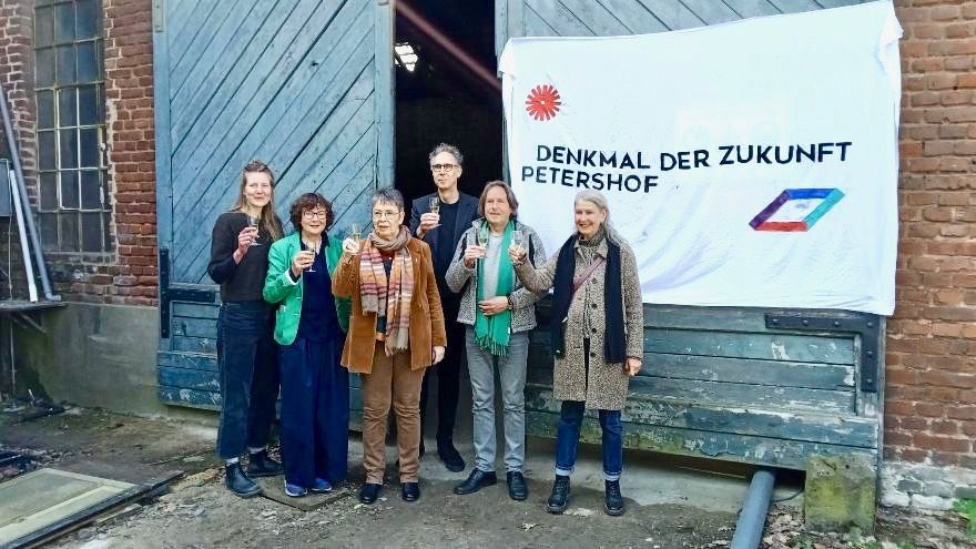 Sechs Menschen stehen mit Sektgläsern in der Hand vor einem alten Gemäuer, an dem ein Transparent hängt: Denkmal der Zukunft, Petershof