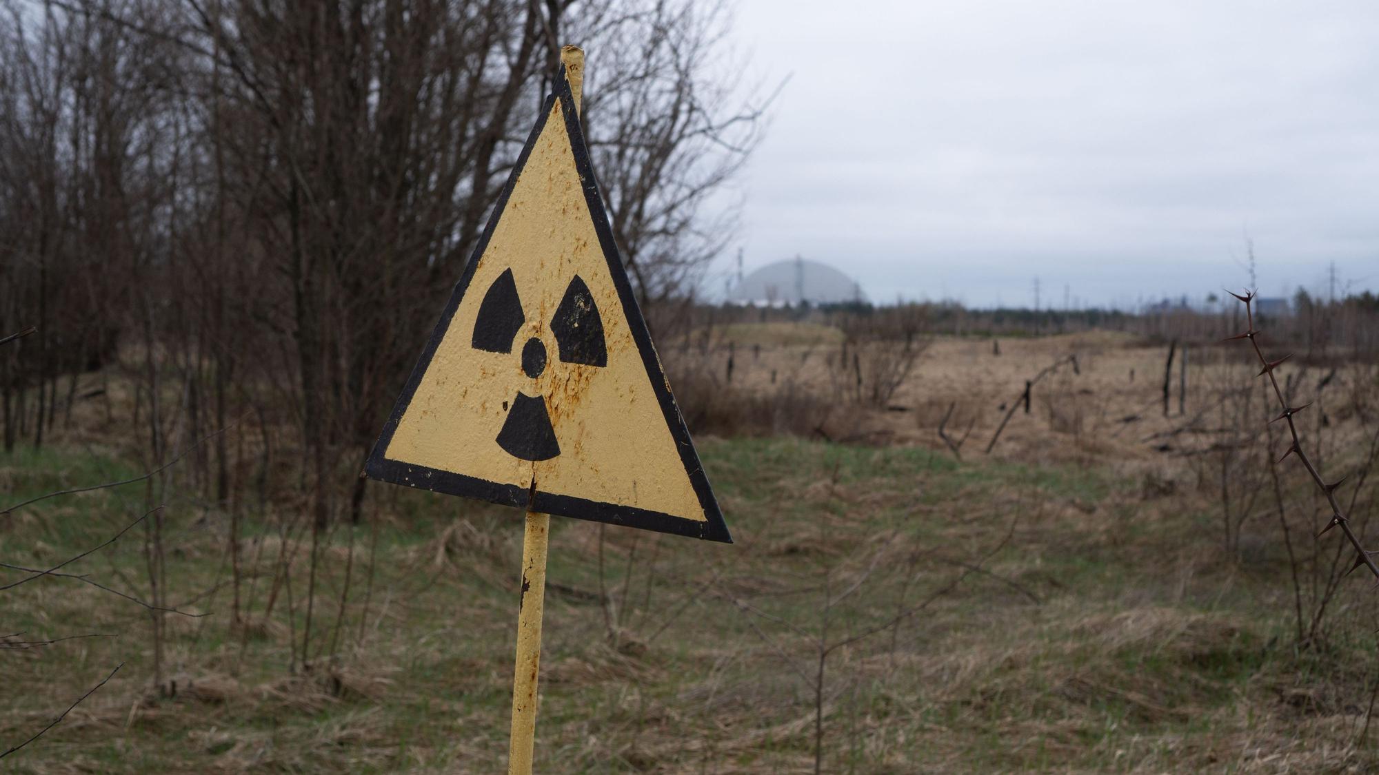 09.04.2026, Ukraine, Tschernobyl: Ein Radioaktivitäts-Warnzeichen, im Hintergrund das Kernkraftwerk Tschernobyl, aufgenommen am 9. April 2026. Was als harmloser Sicherheitstest begann, entwickelte sich am 26. April 1986 zur bedeutendsten Atomkatastrophe der Geschichte. An diesem Tag explodierte Reaktor 4 des Wladimir-Iljitsch-Lenin-Kernkraftwerks, weitläufig bekannt als Kernkraftwerk Tschernobyl. Foto: Nina Mesitz/APA/dpa +++ dpa-Bildfunk +++