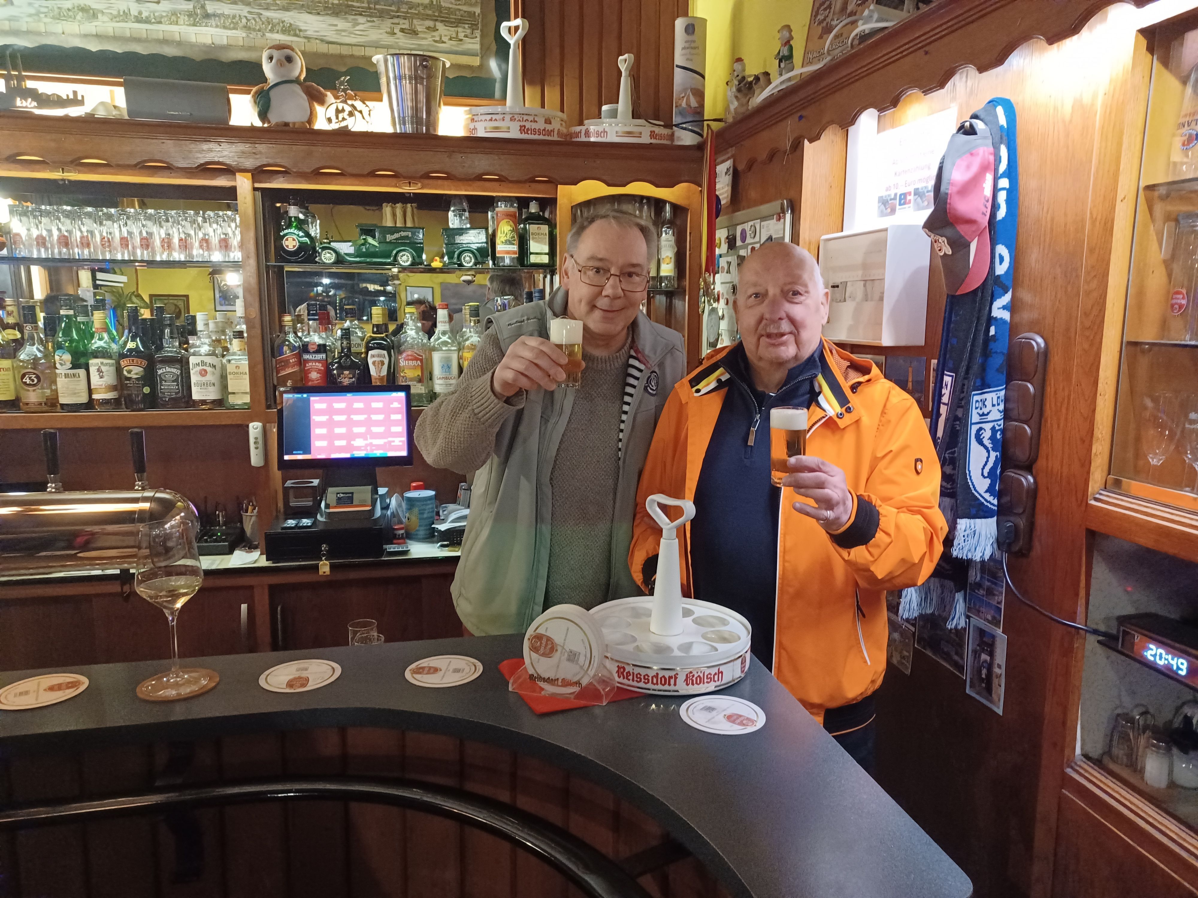 Zwei Männer stehen hinter einer Theke in einer Kneipe und prosten mit einem Kölsch.