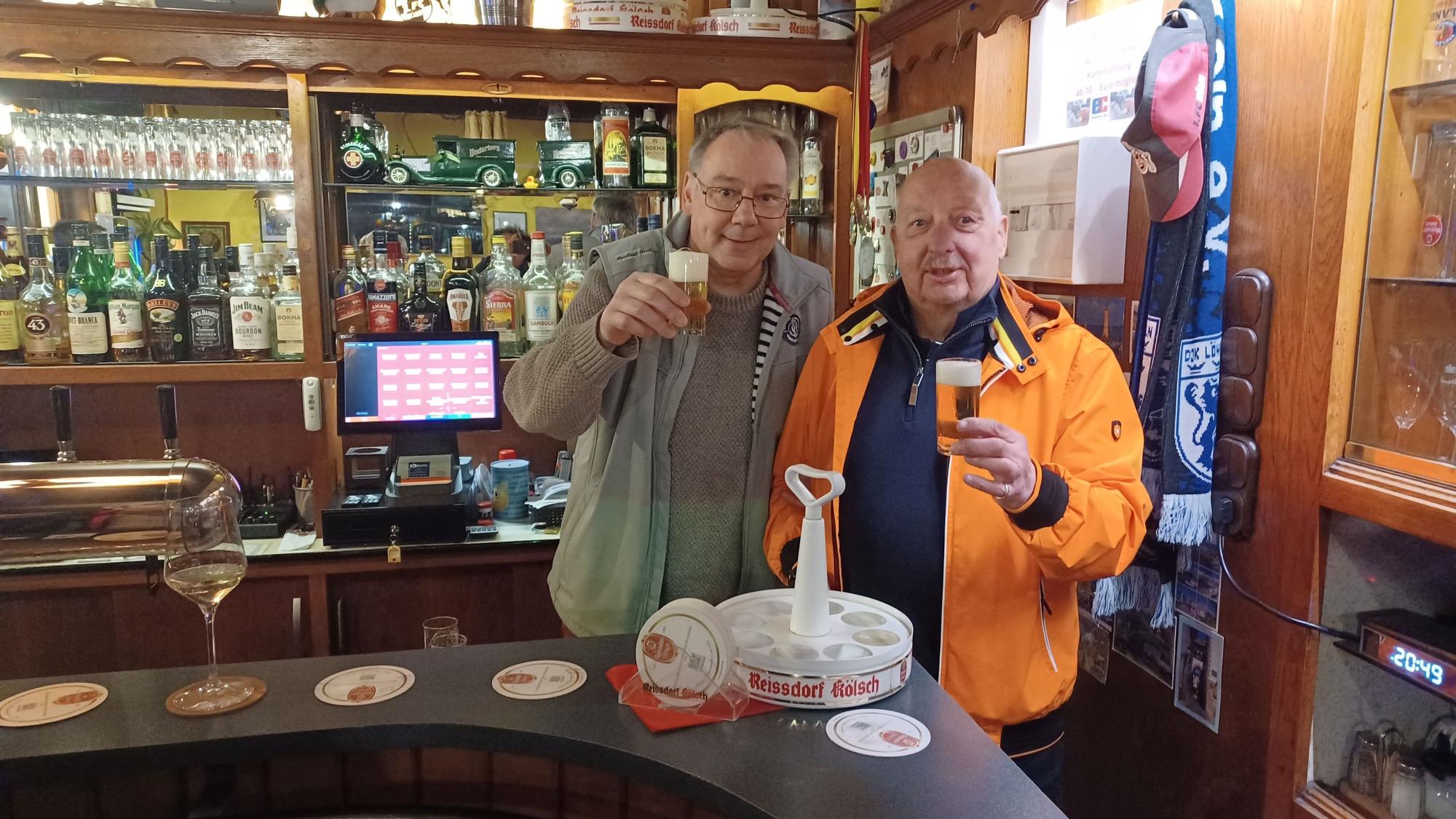 Zwei Männer stehen hinter einer Theke in einer Kneipe und prosten mit einem Kölsch.