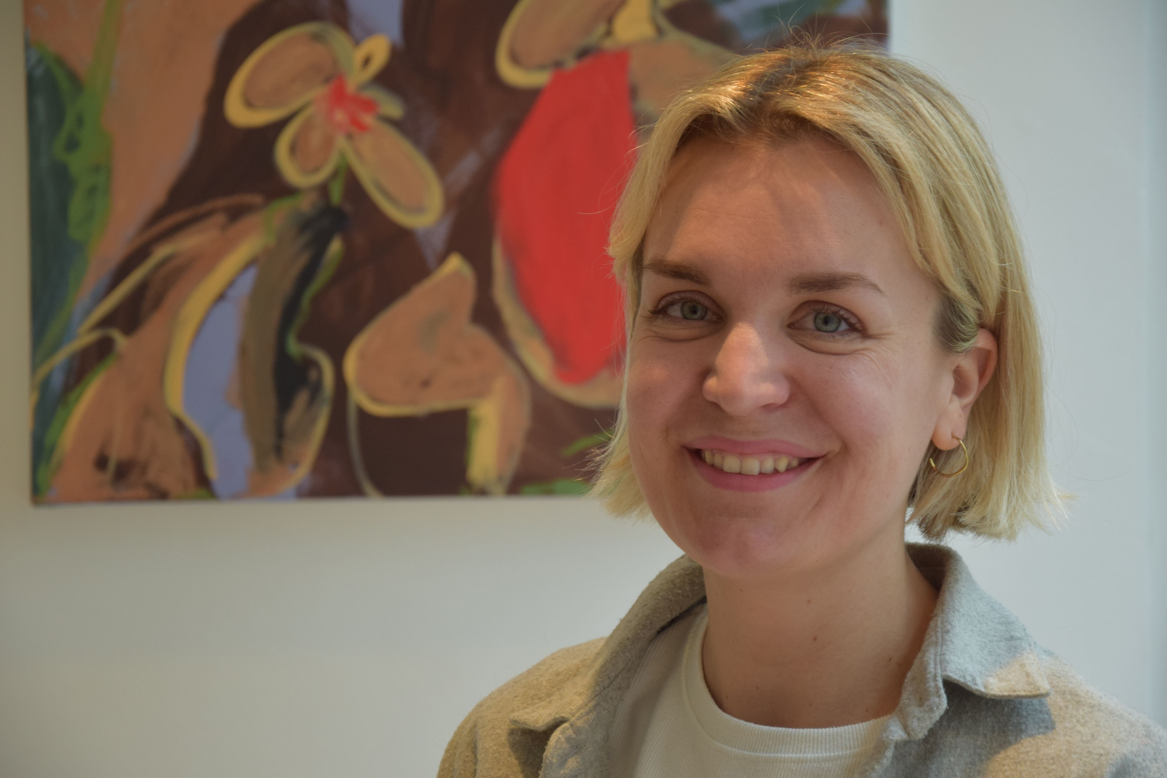 Eine Frau mit blonden, kurzen Haaren steht vor einer weißen Wand, an der ein buntes Gemälde hängt.