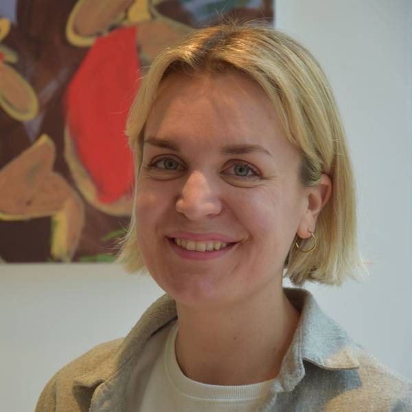 Eine Frau mit blonden, kurzen Haaren steht vor einer weißen Wand, an der ein buntes Gemälde hängt.