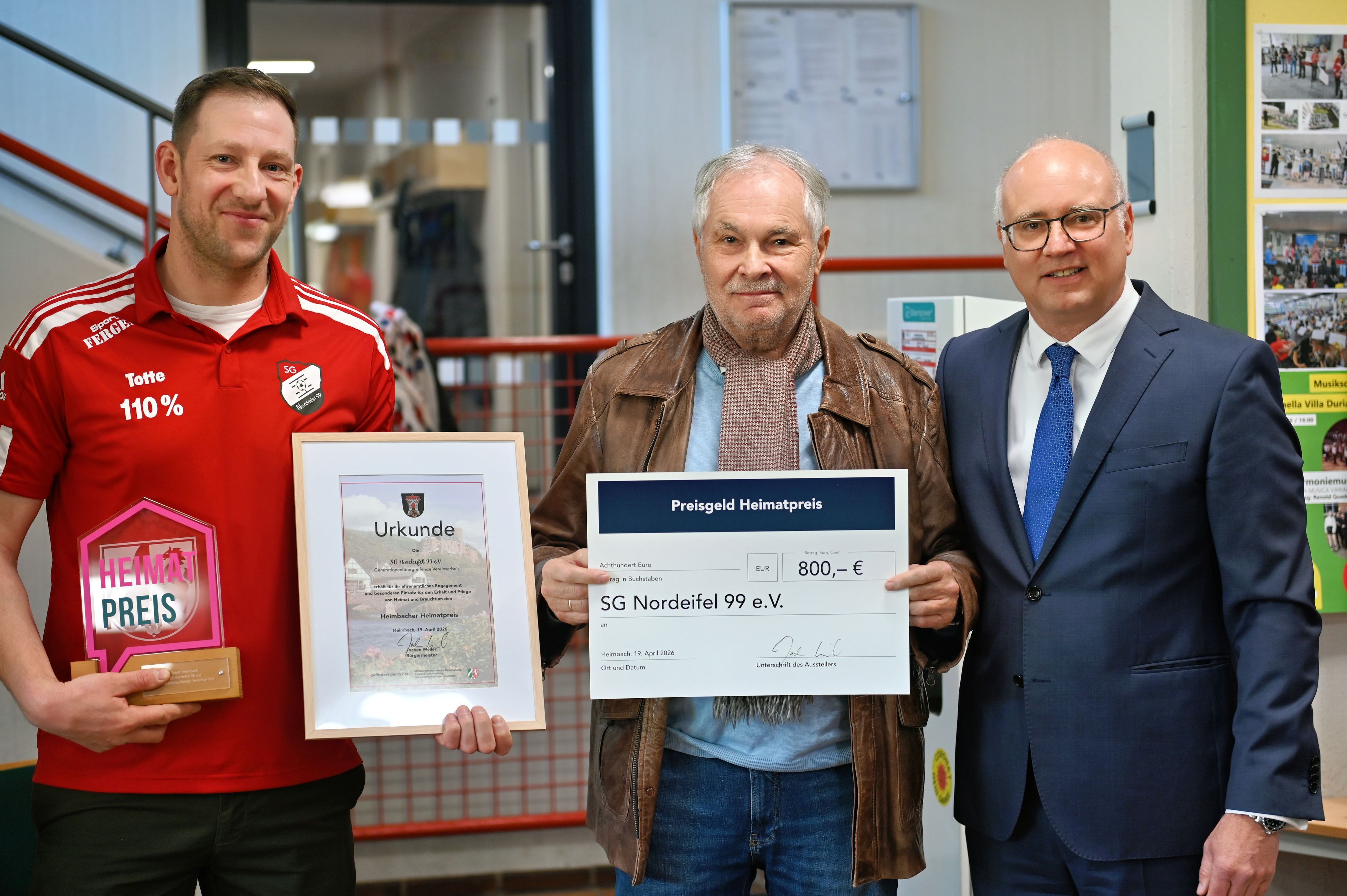 Zwei Männer schauen in die Kamera, der linke rägt ein rotes Trikot, der rechte eine braune Lederjacke.