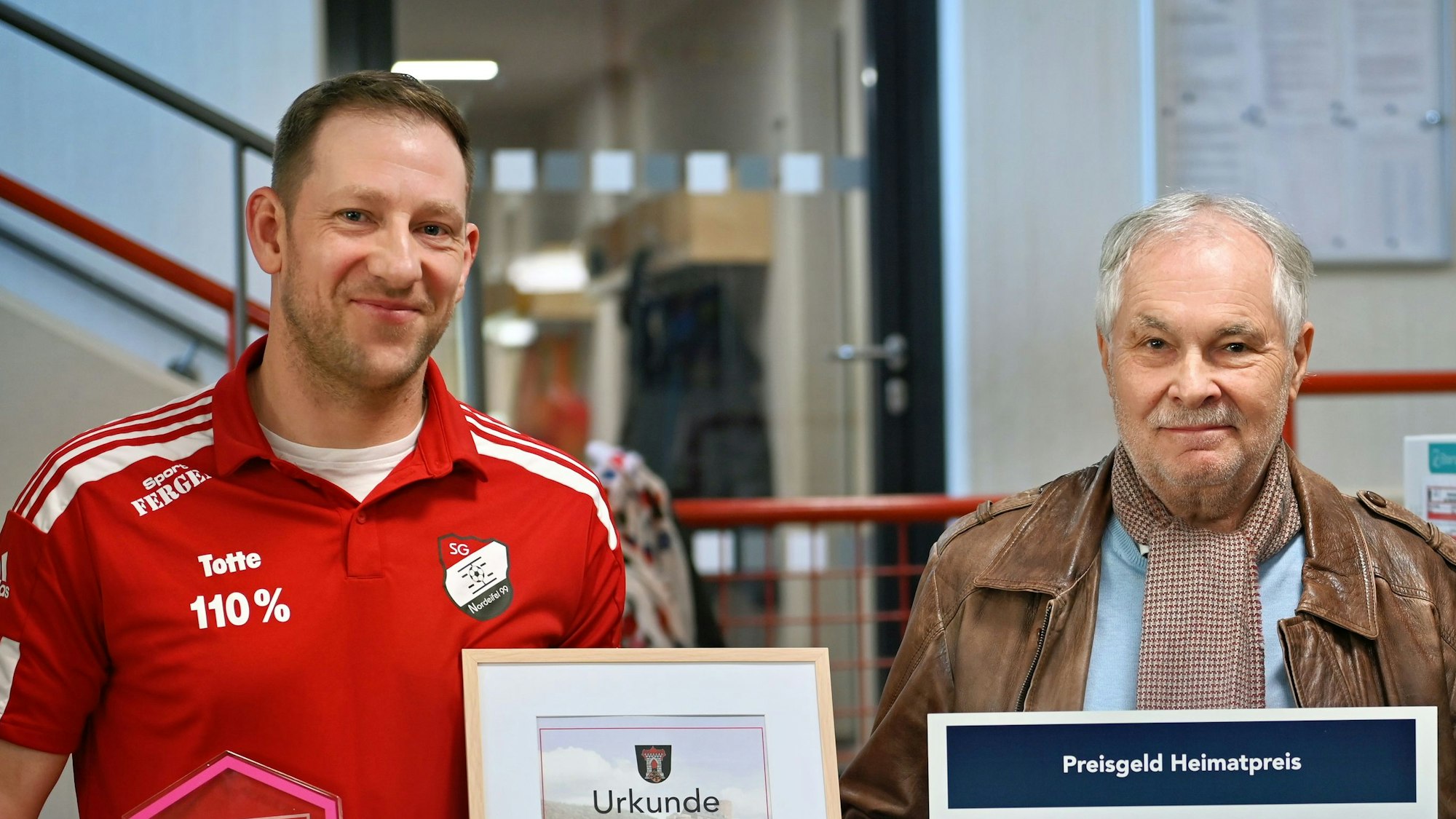 Zwei Männer schauen in die Kamera, der linke rägt ein rotes Trikot, der rechte eine braune Lederjacke.
