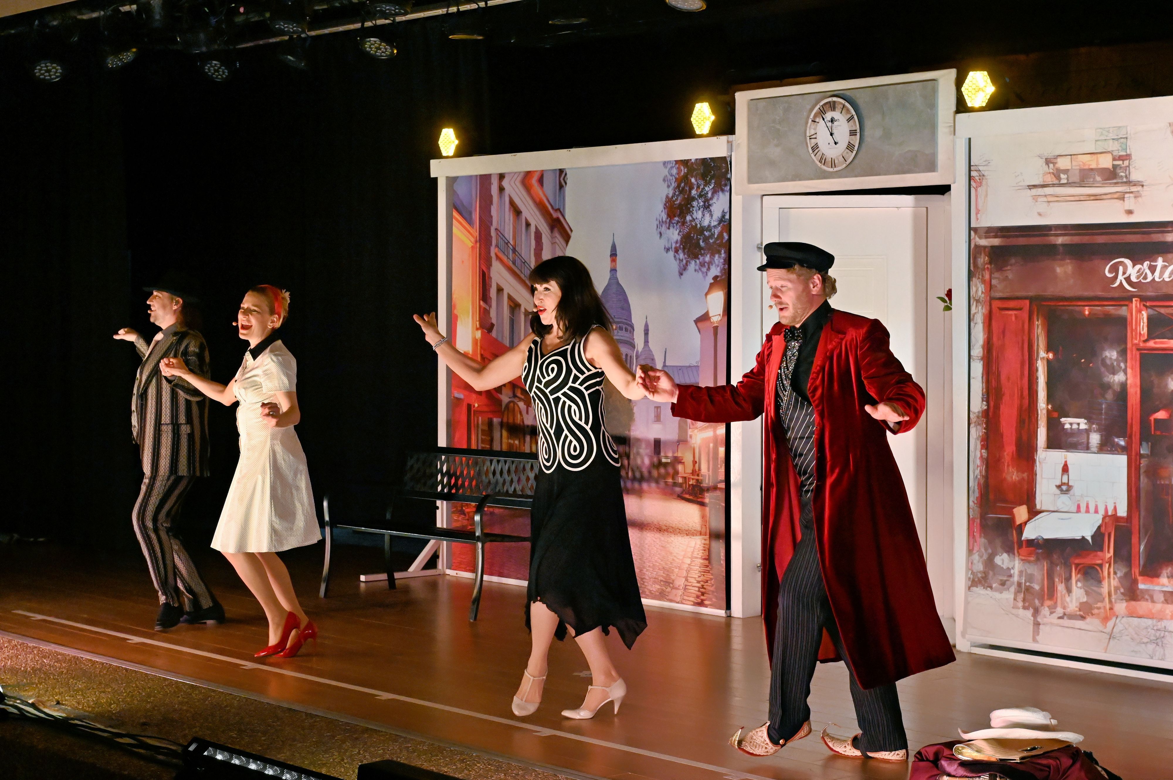 Zwei Männer und zwei Frauen tanzen vor einem Bühnenbild, das eine Pariser Straße darstellt.