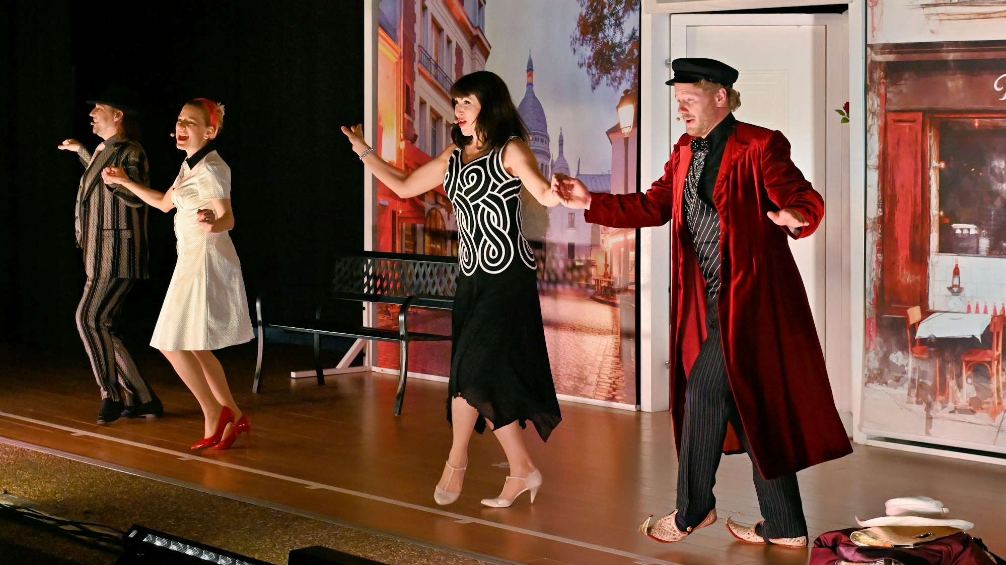 Zwei Männer und zwei Frauen tanzen vor einem Bühnenbild, das eine Pariser Straße darstellt.