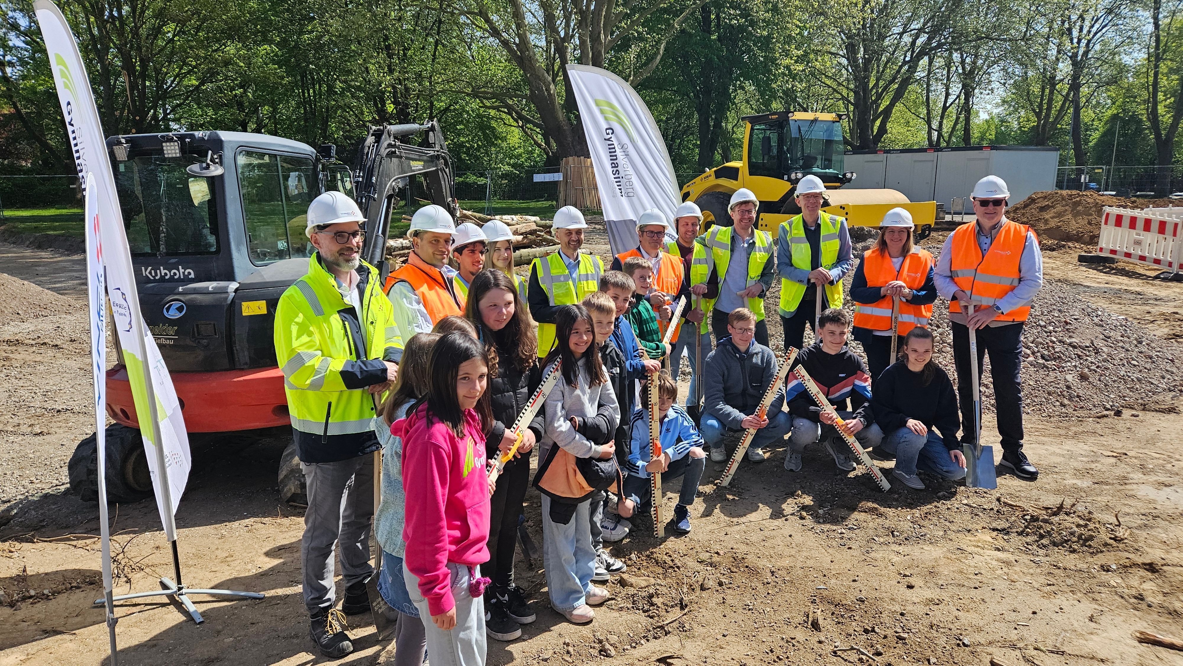Zu sehen sind Menschen mit Helmen und Warnwesten sowie Schülerinnen und Schüler auf einer Baustelle.