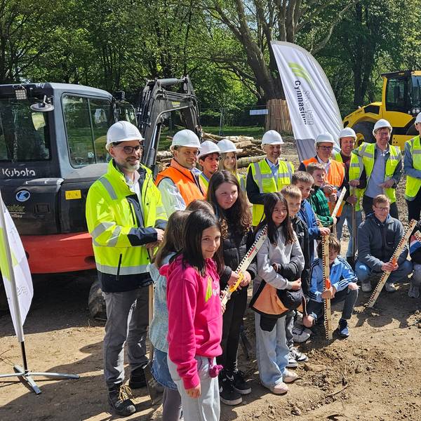 Zu sehen sind Menschen mit Helmen und Warnwesten sowie Schülerinnen und Schüler auf einer Baustelle.