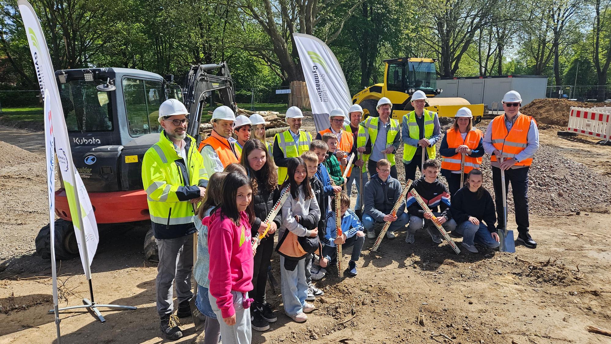 Zu sehen sind Menschen mit Helmen und Warnwesten sowie Schülerinnen und Schüler auf einer Baustelle.