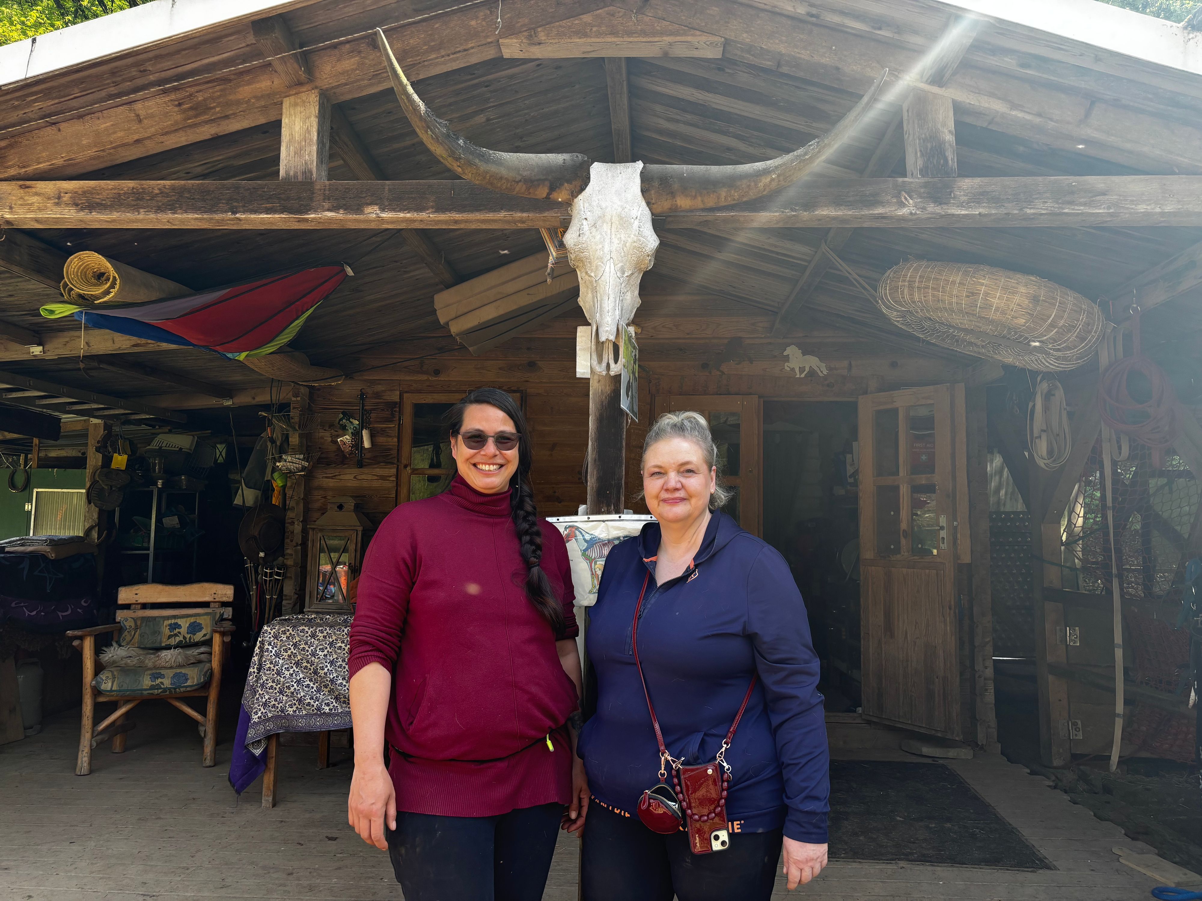Zwei Frauen stehen nebeneinander und blicken lächelnd in die Kamera. Im Hintergrund ist die Ranch zu sehen.