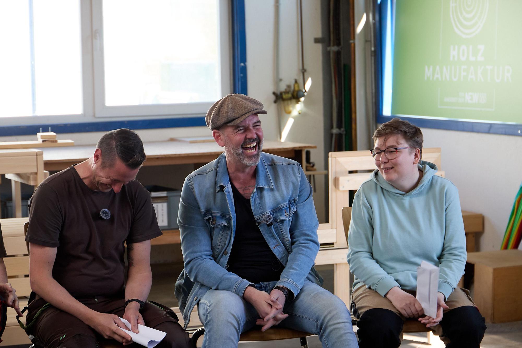 Doc Esser sitzt zwischen einem Mann und einer Frau in Arbeitskleidung und lacht herzlich.