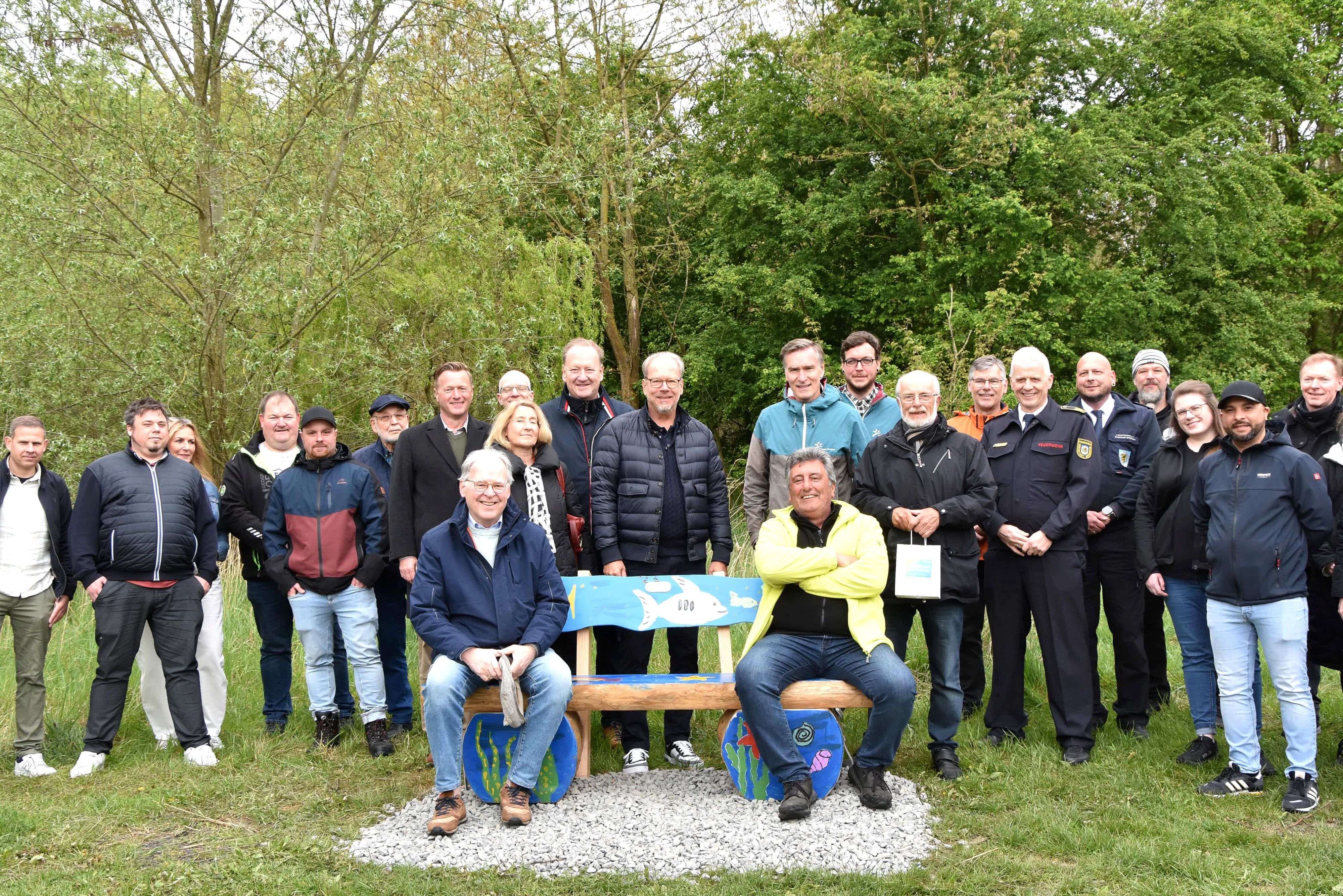 Zwei Männer sitzen auf einer Bank, hinter stehen Frauen und Männer.