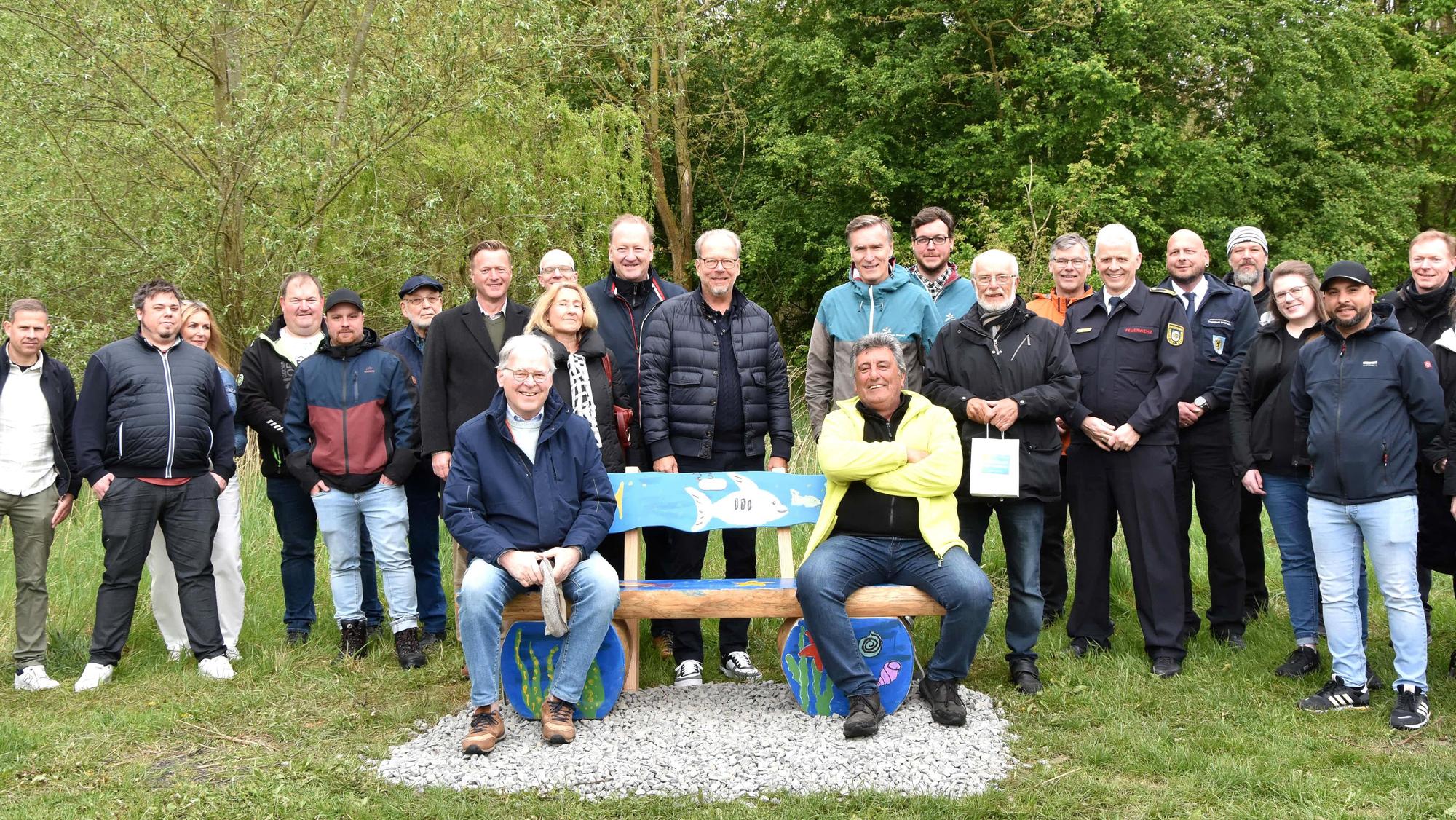 Zwei Männer sitzen auf einer Bank, hinter stehen Frauen und Männer.