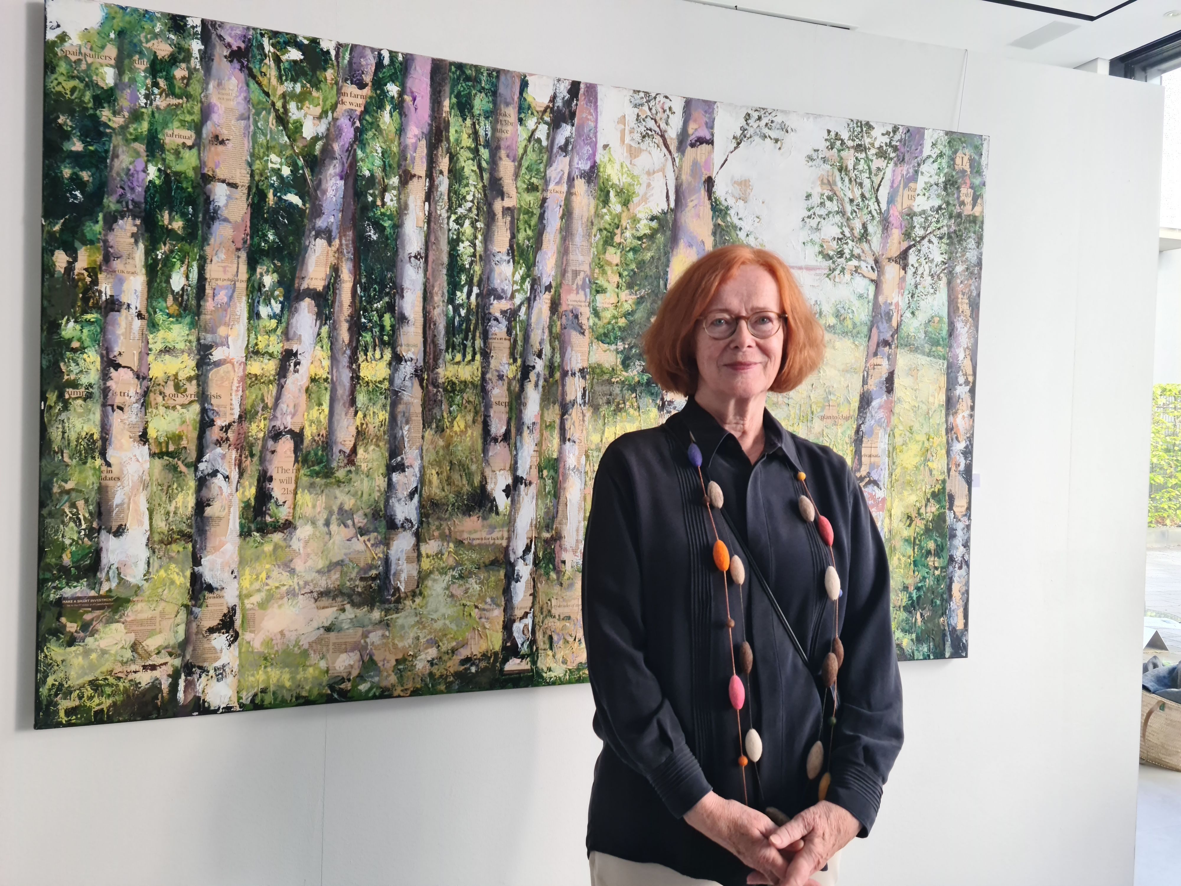 Eine Frau mit rotem Haar und Brille steht vor dem Bild eines Birkenwalds.