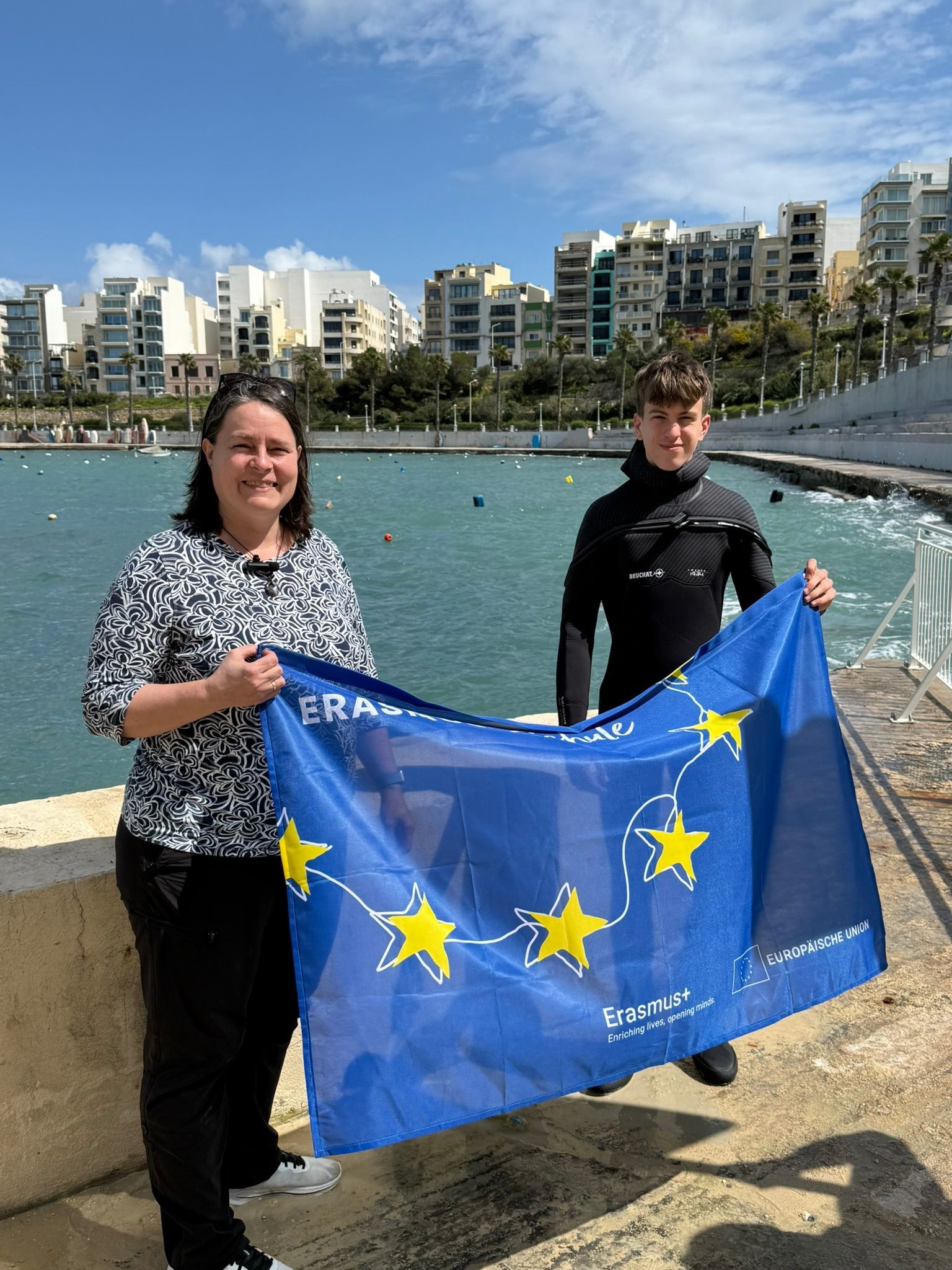 Eine Dame mittleren Alters und ein Schüler stehen am Meer und halten eine blaue Fahne in der Hand.