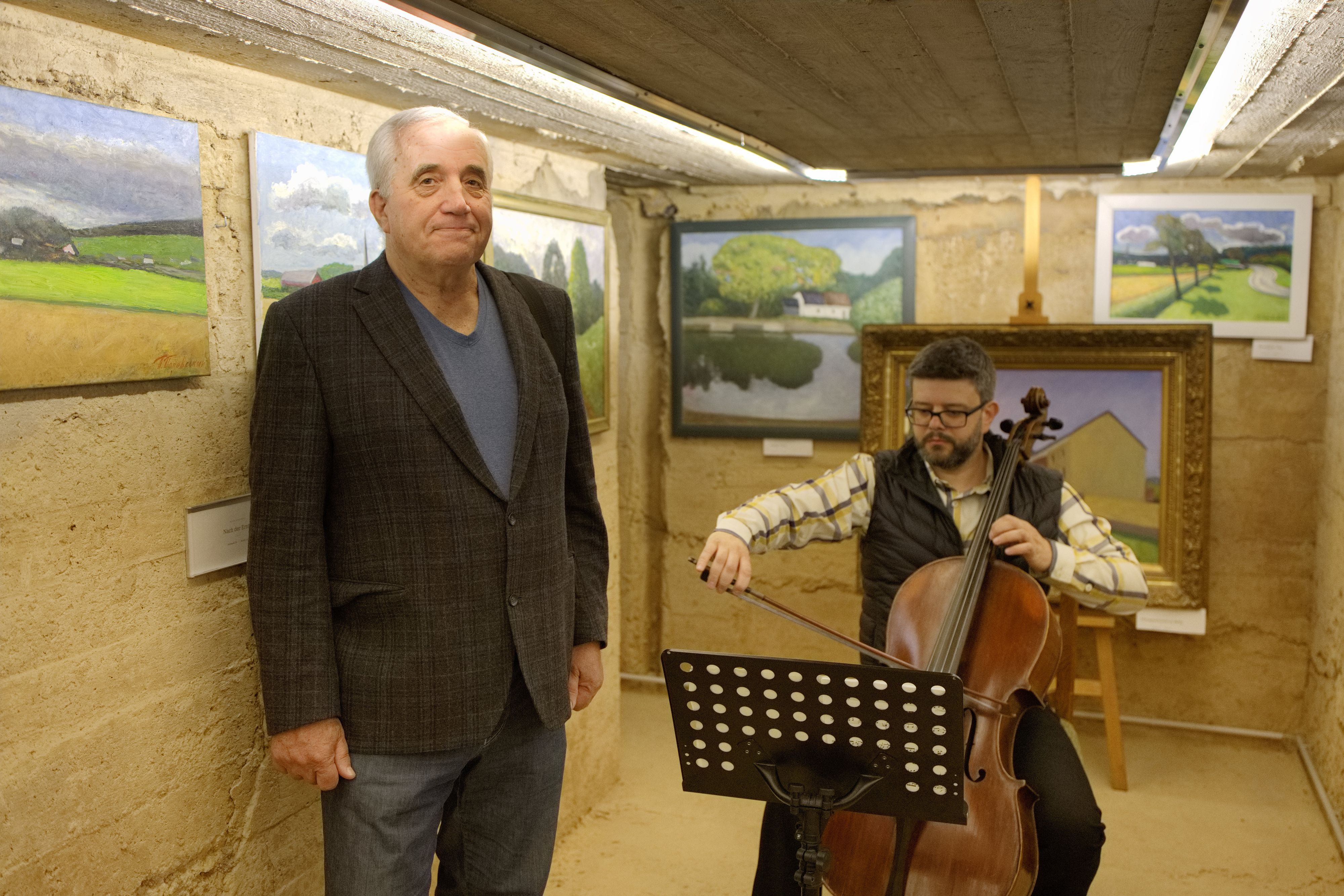 Ein älterer Mann steht vor Bildern, während neben ihm ein jüngerer Mann auf einem Bass musiziert.