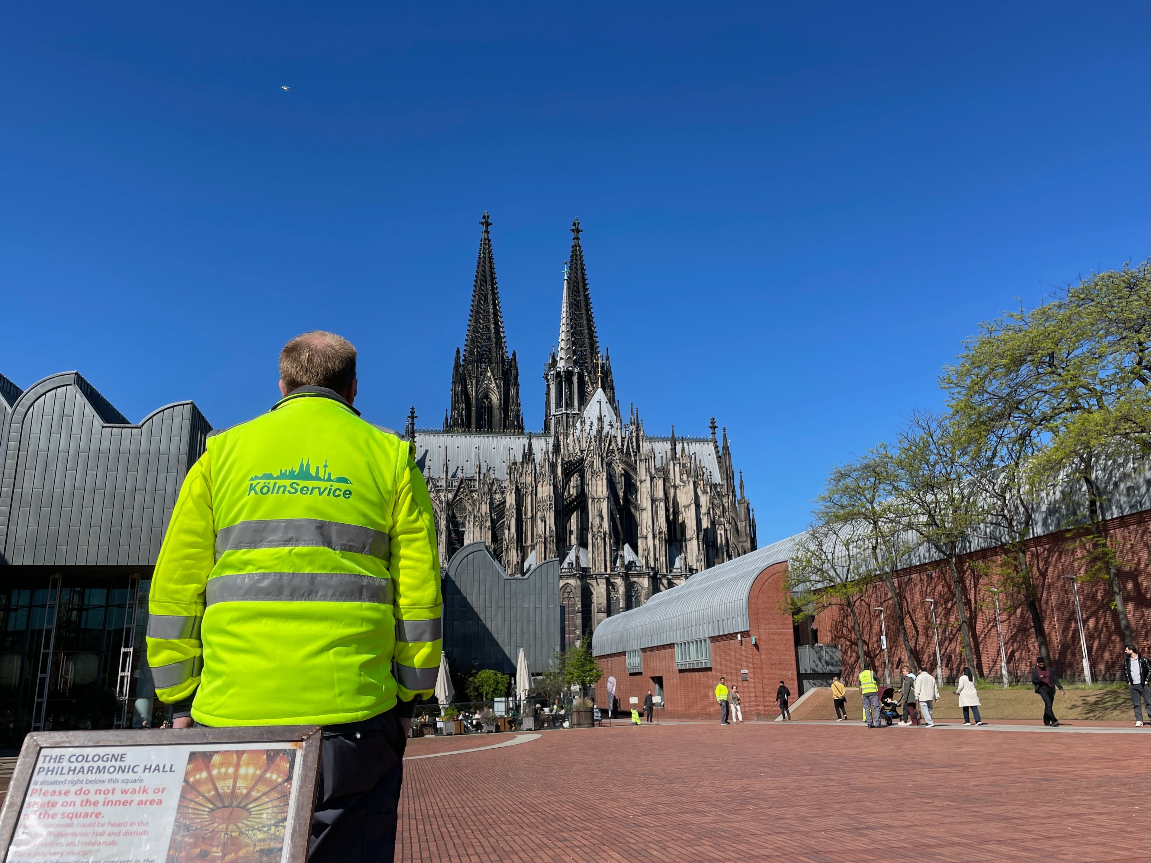Ein Mann in gelber Warnweste steht auf einem Platz, im Hintergrund sind das Museum Ludwig und der Dom zu sehen.