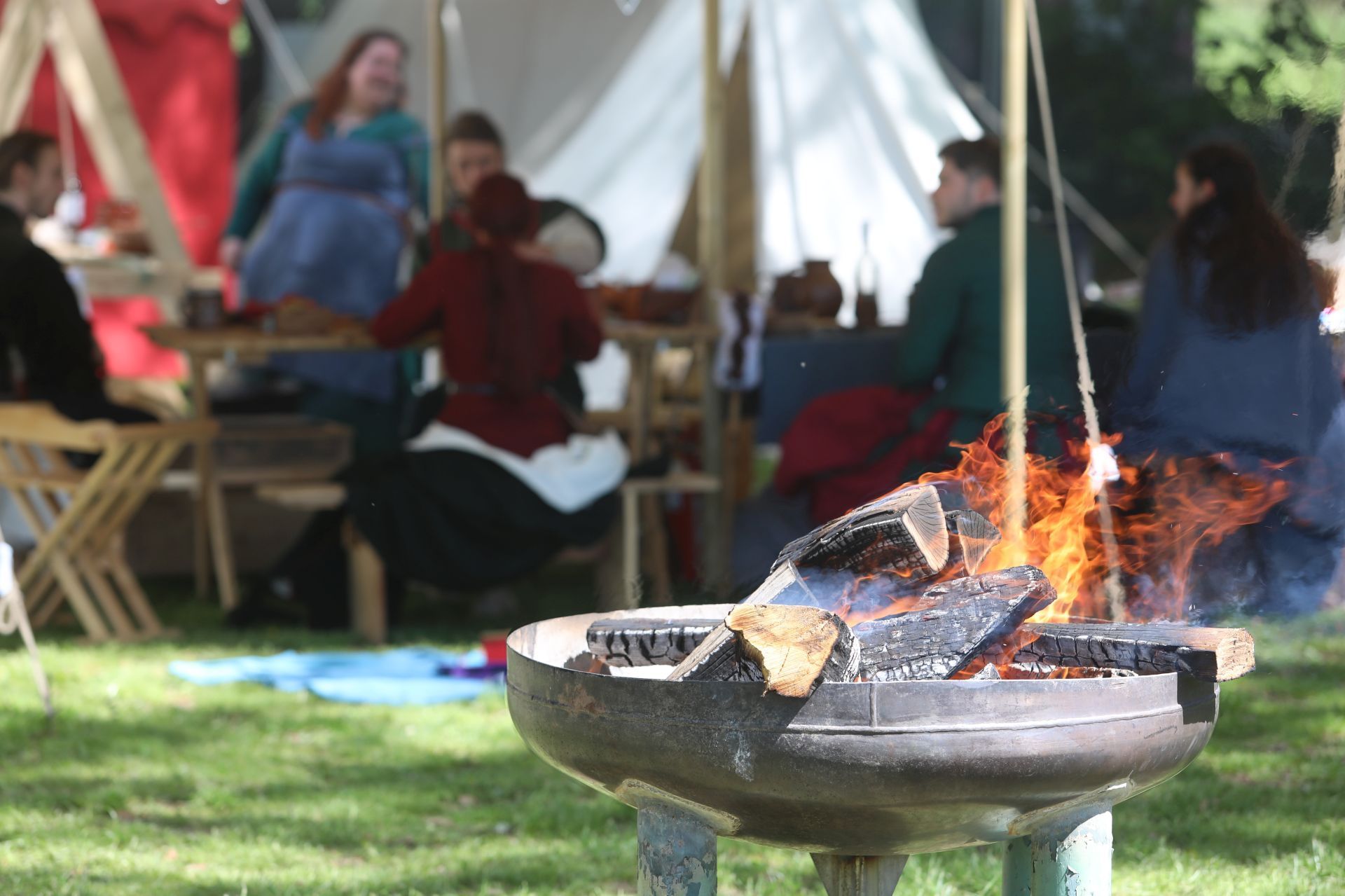 Eine Feuerschale mit brennenden Holzscheiten, im Hintergrund Menschen vor Zelten.