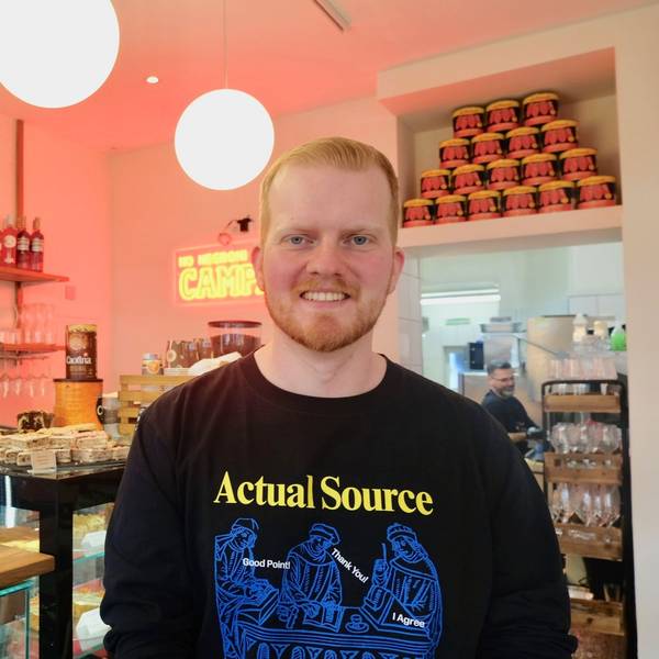 Ein junger Mann mit kurzem, rotblonden Haar und Bart steht vor einer Kuchentheke in einem Café.