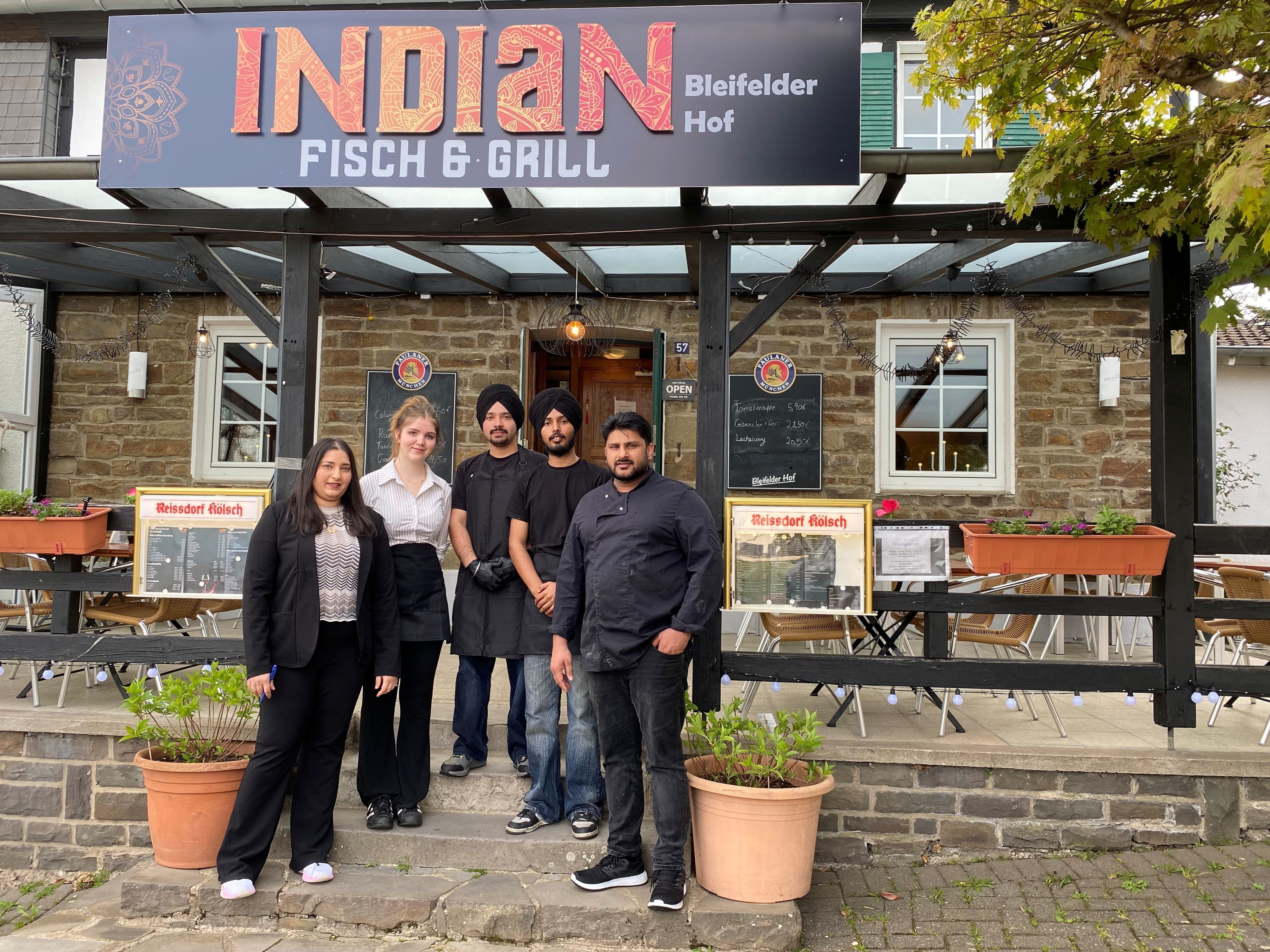 Oben das Schild des Restaurants mit seinem Namen, davor die beiden Inhaber, eine junge Frau aus Bleifeld und zwei junge Männer aus Indien, beide mit schwarzem Turban.
