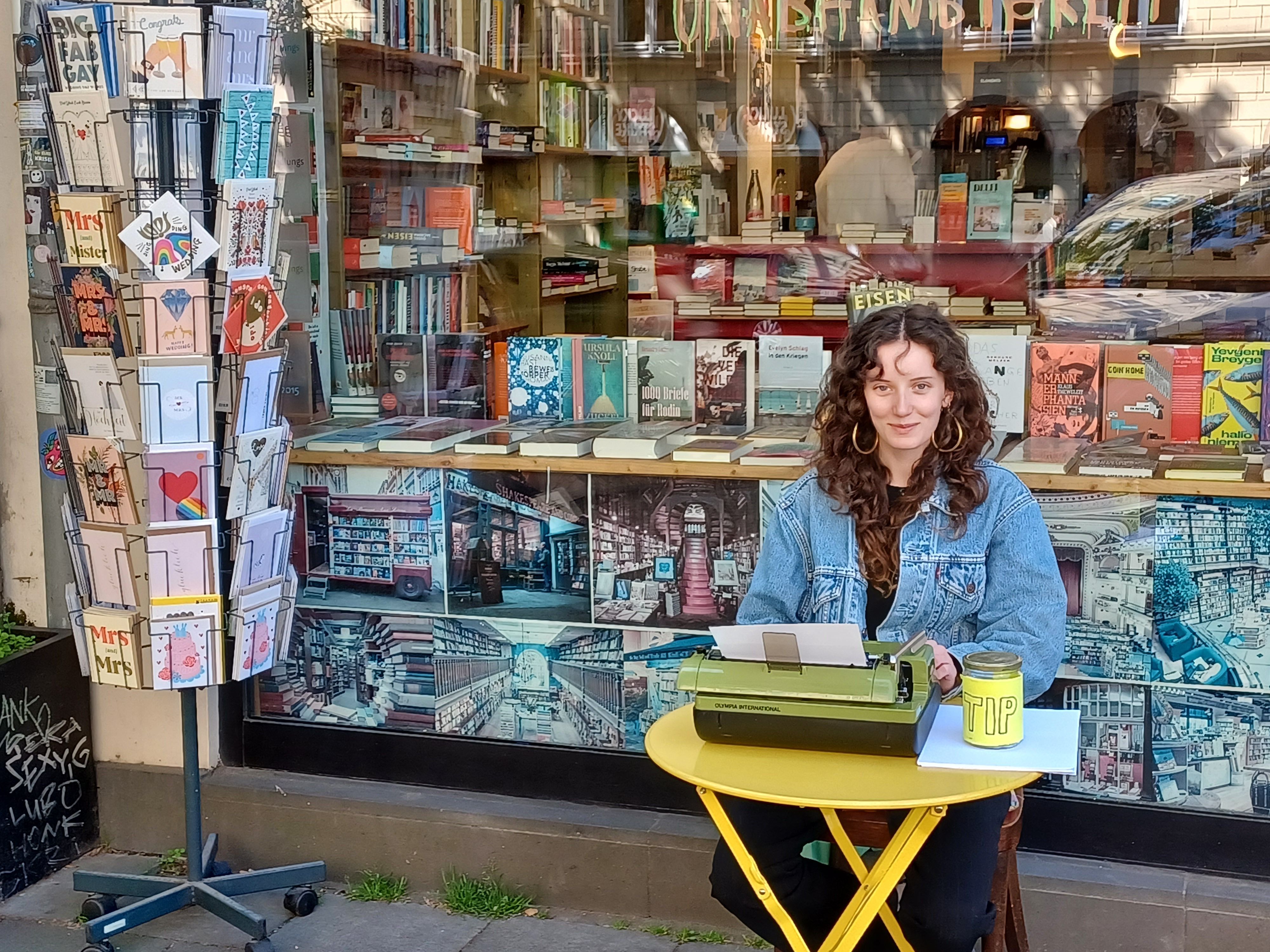 Eine junge Frau mit lockigem Haar sitzt an einem kleinen gelben Tisch vor einer Buchhandlung. Sie schreibt auf einer grünen Schreibmaschine.